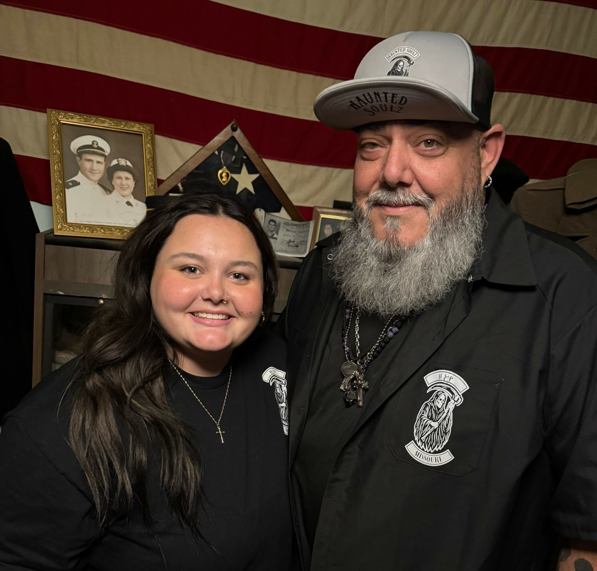 A young woman and a man with a beard pose together indoors, with a framed photo and American flag in the background.