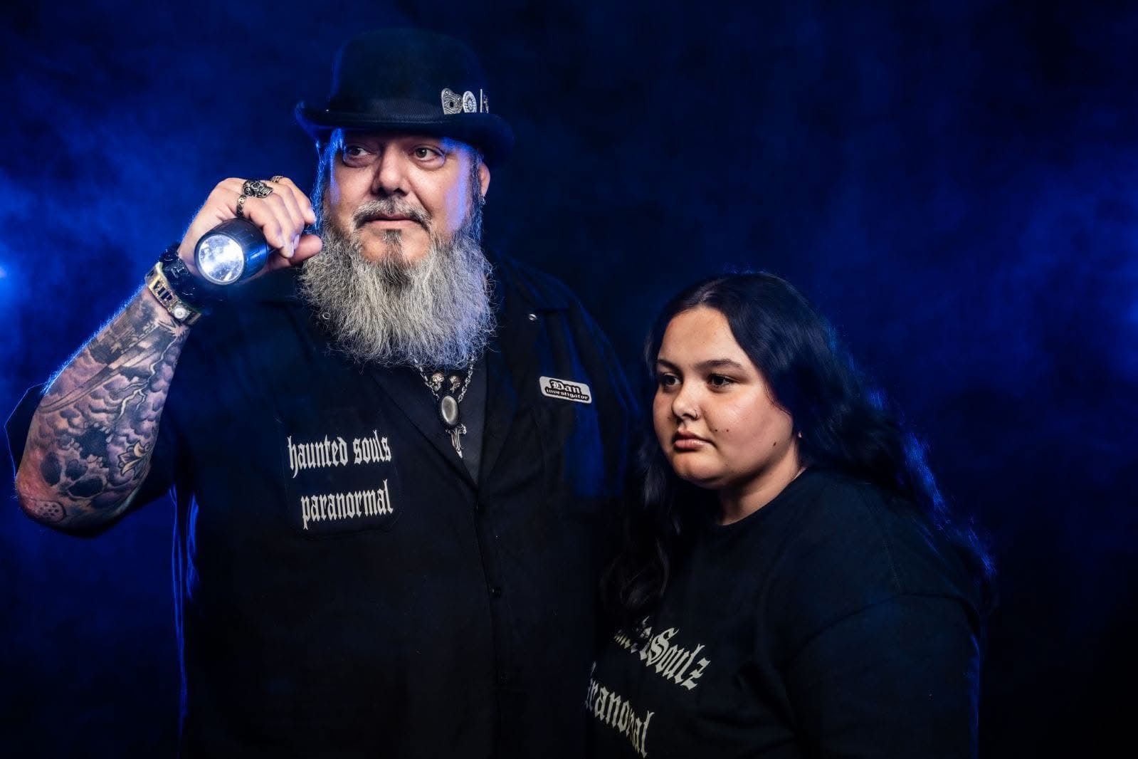 A bearded man with tattoos and a woman stand together against a dark background, both wearing black clothing.