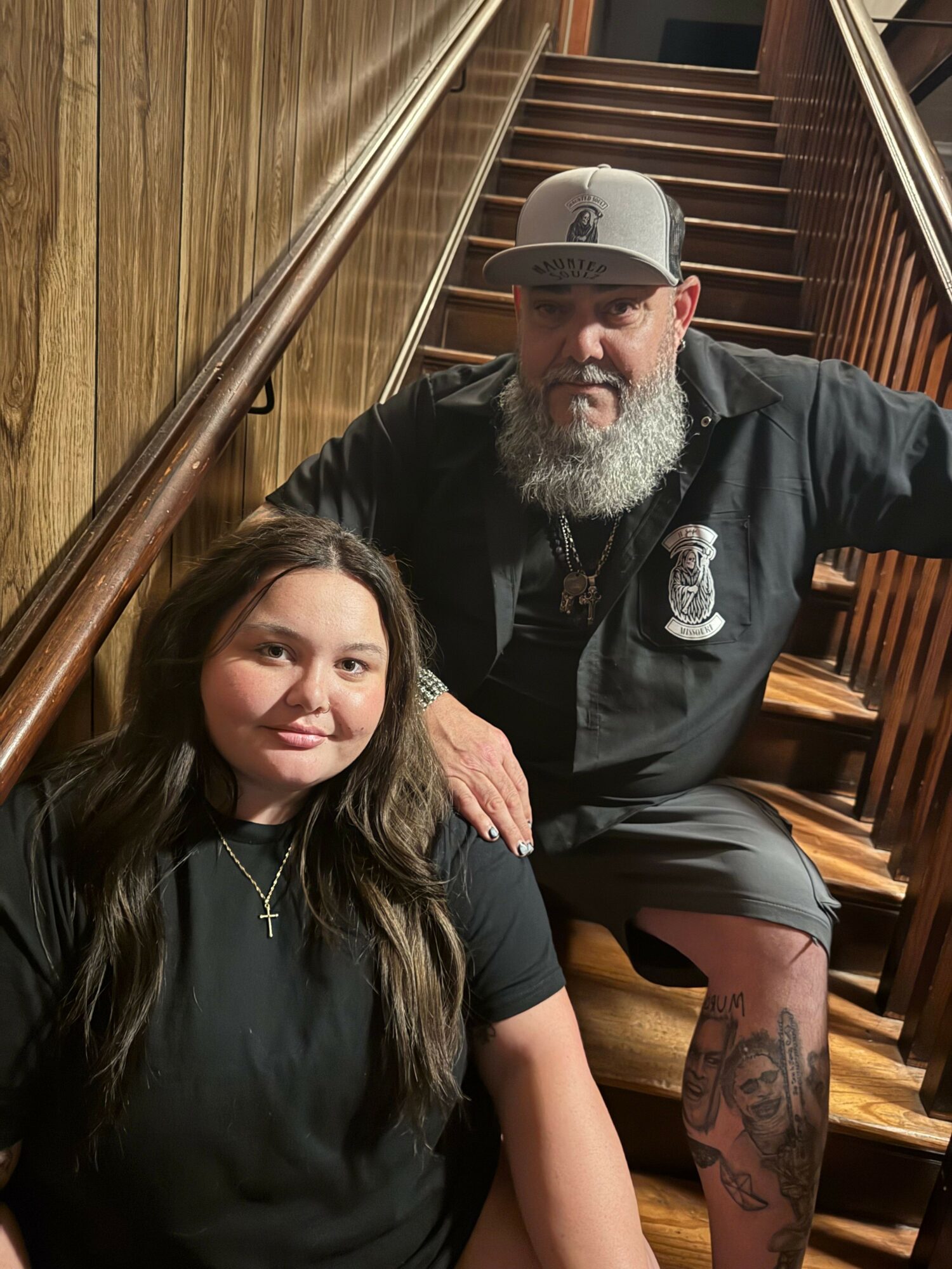 A man with a beard and tattoos, wearing a cap and black jacket, sitting next to a woman with long dark hair, both on a wooden bench.