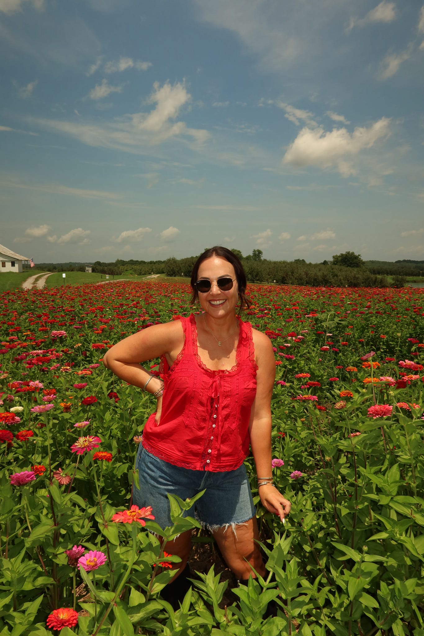 Woman in sunglasses standing in a field of pink and red flowers under a partly cloudy sky.