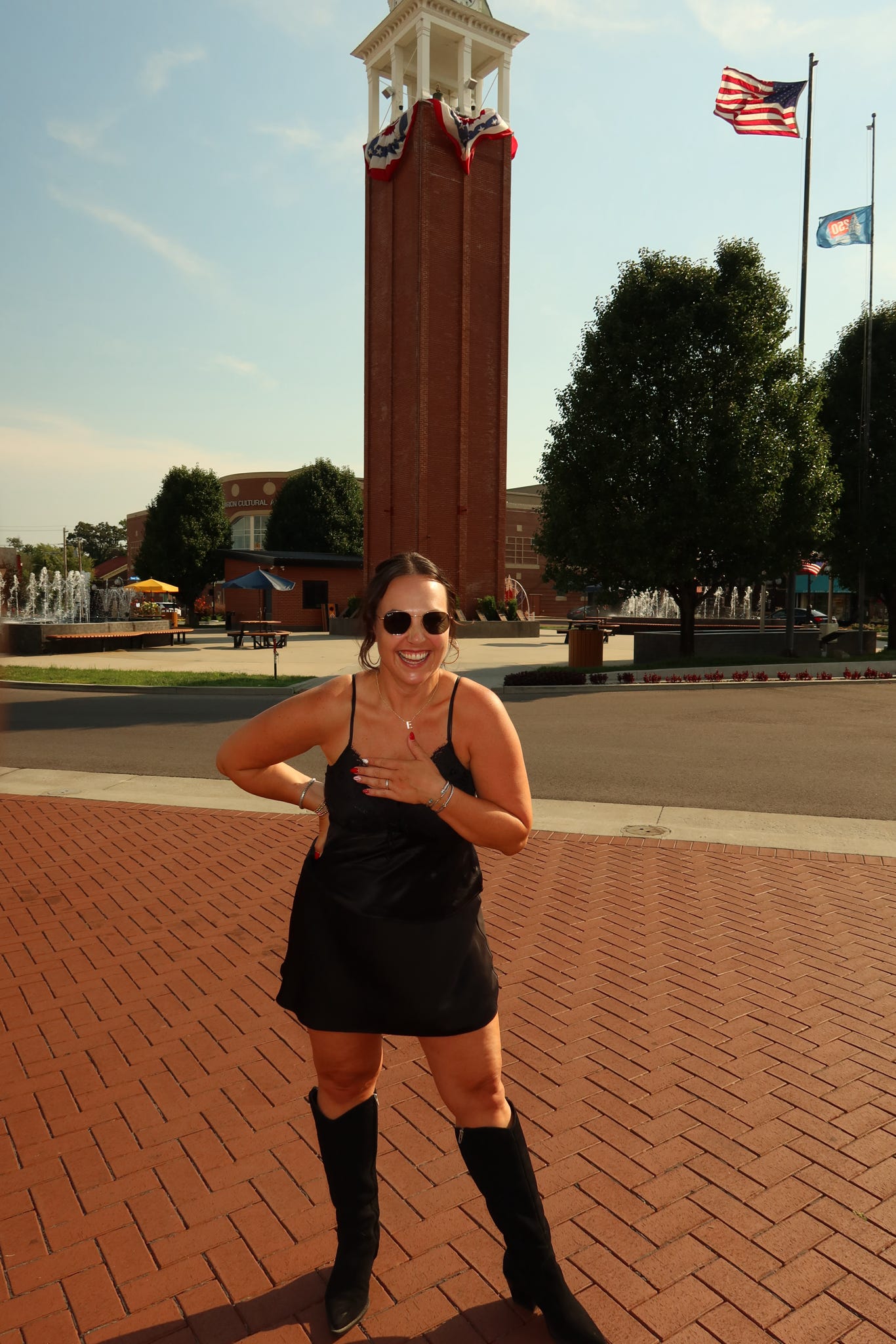 Woman in black dress and sunglasses standing outdoors with a tall clock tower and flags in the background.