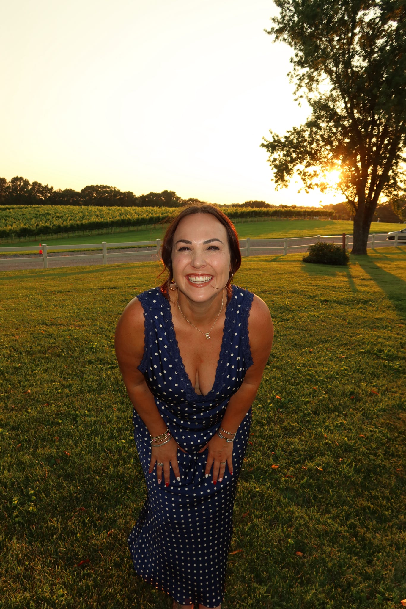 Woman smiling outdoors at sunset, standing on grass with trees and a road in background.