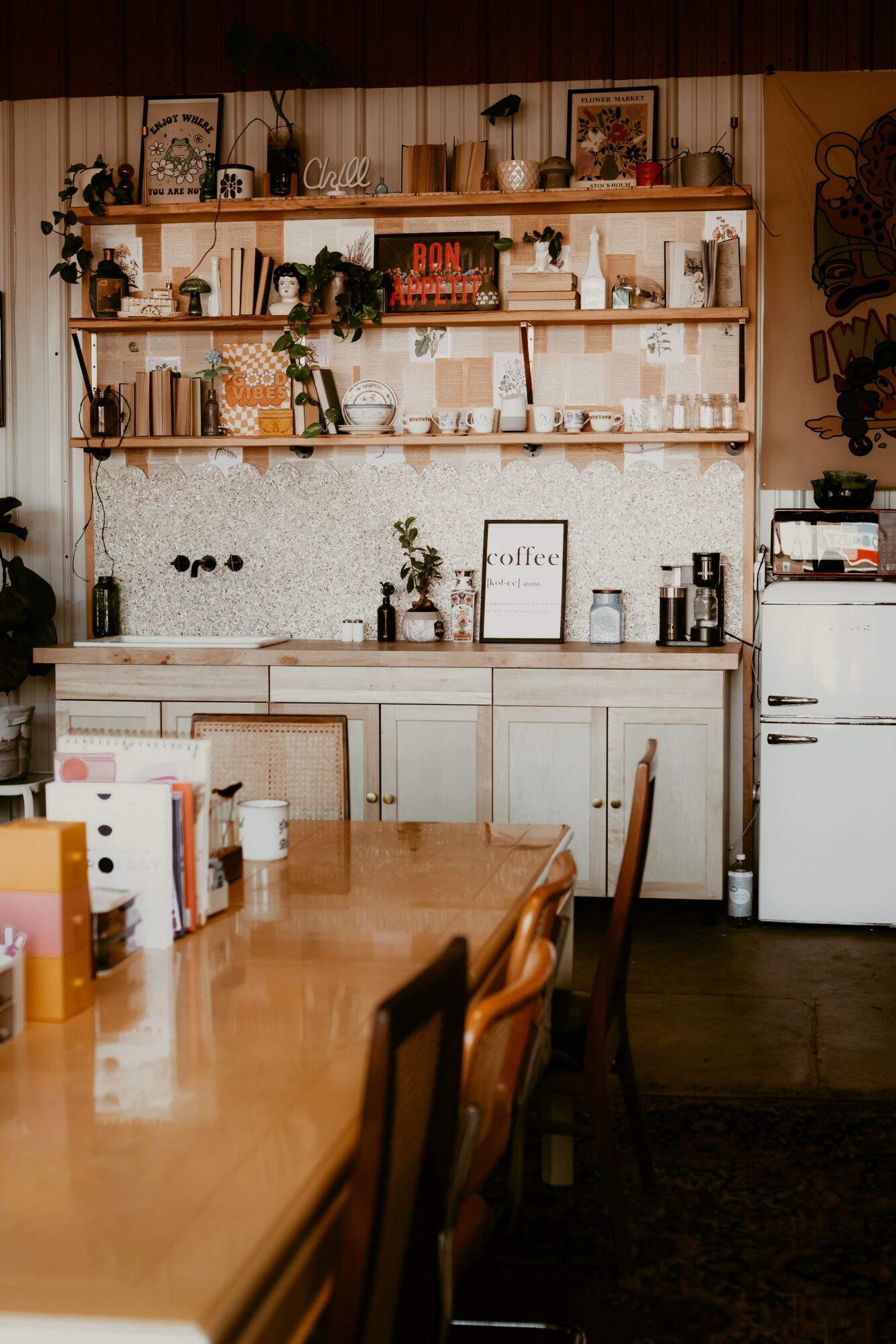 Kitchen with wooden shelves, plants, framed pictures, and a white refrigerator, viewed from a dining table with chairs.