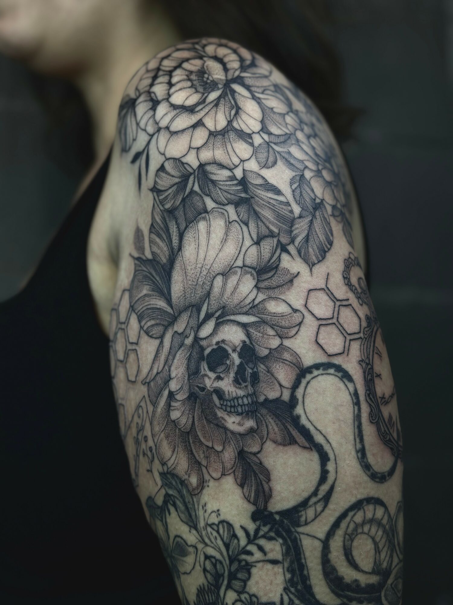 Tattoo of flowers, a skull, and a snake on a person's upper arm and shoulder, in black and gray ink.