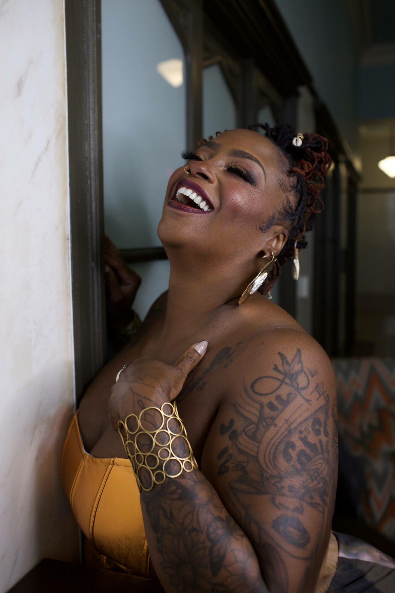 Smiling woman with tattoos and jewelry, leaning against a wall, wearing a yellow top, in a relaxed pose.