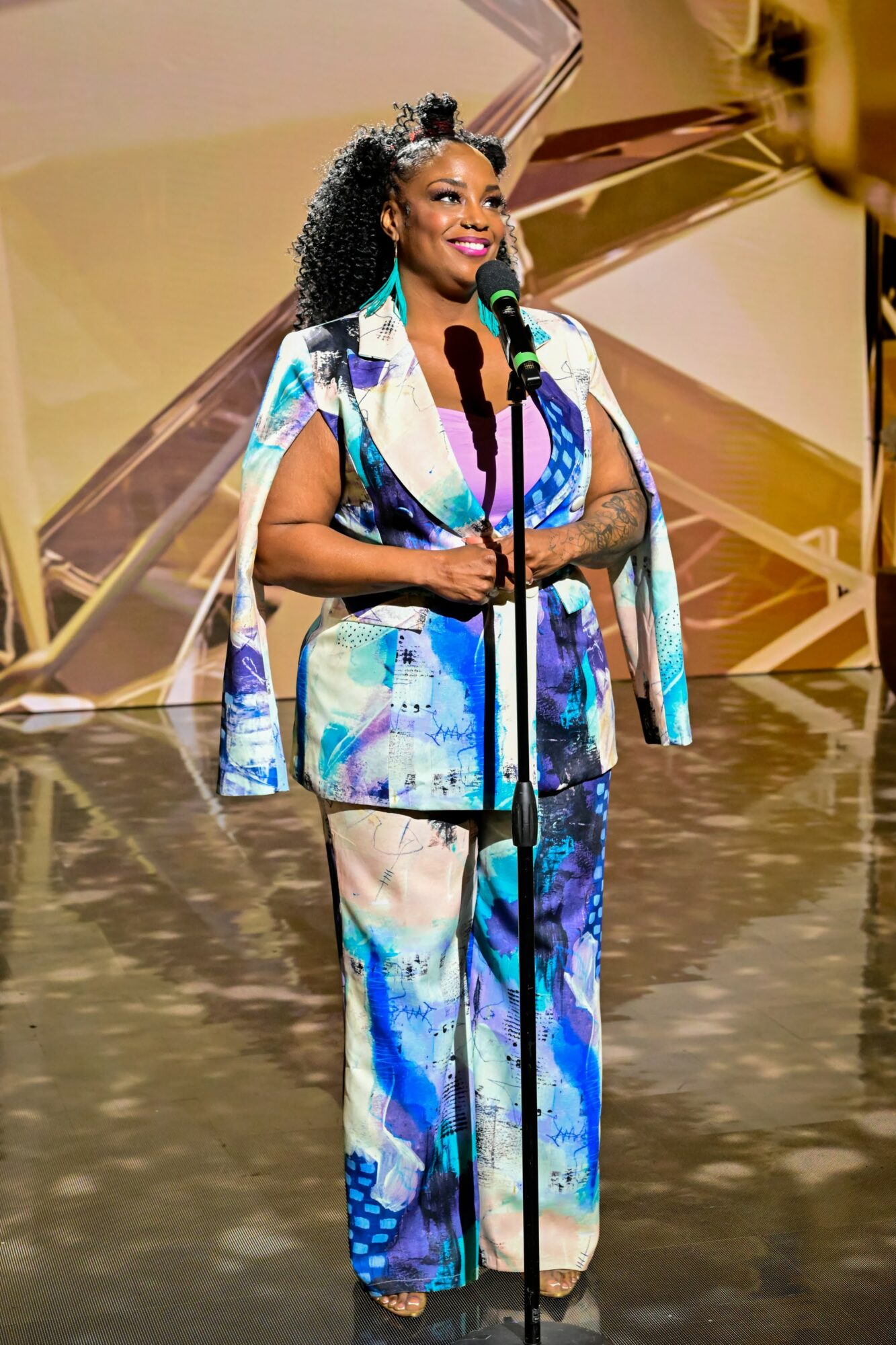 Woman in colorful outfit speaking into a microphone on stage with a gold backdrop.