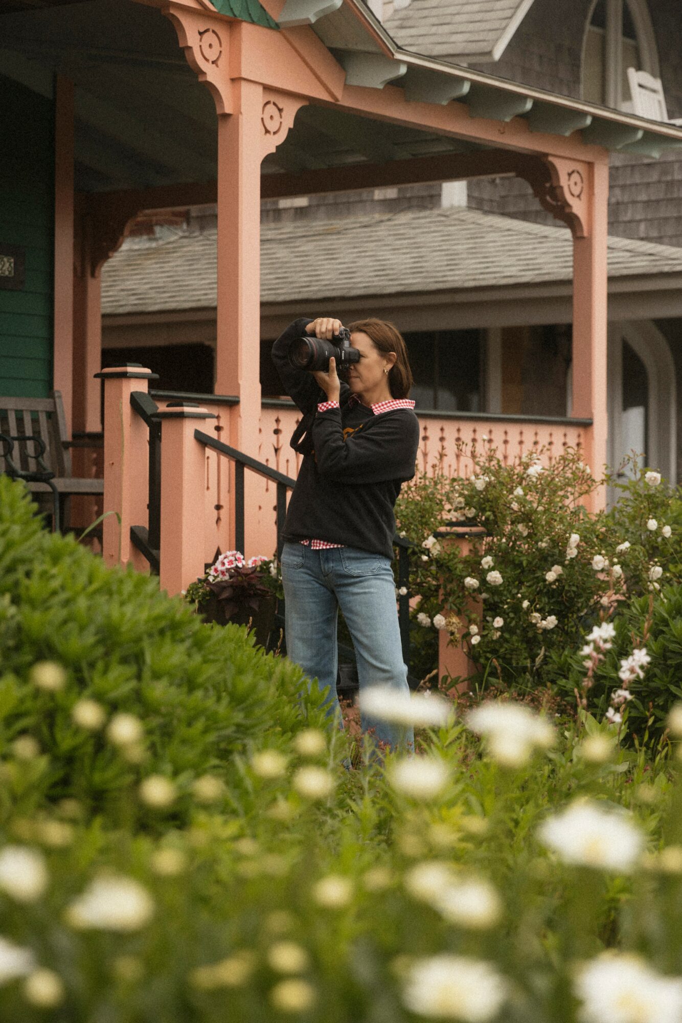 Two women embrace on a porch with pink railing, surrounded by white flowers and greenery, in front of a house.