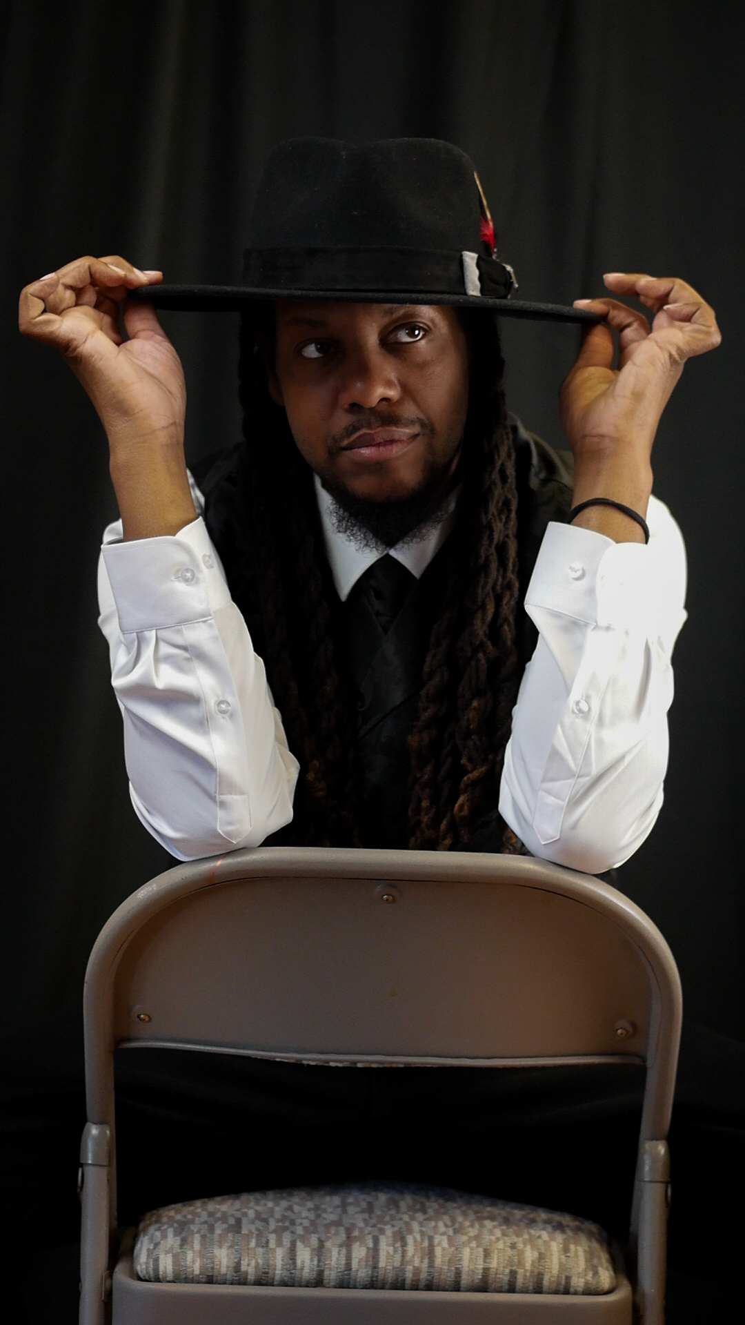 Person with long hair and a hat, holding the brim with both hands, sitting in front of a black curtain.