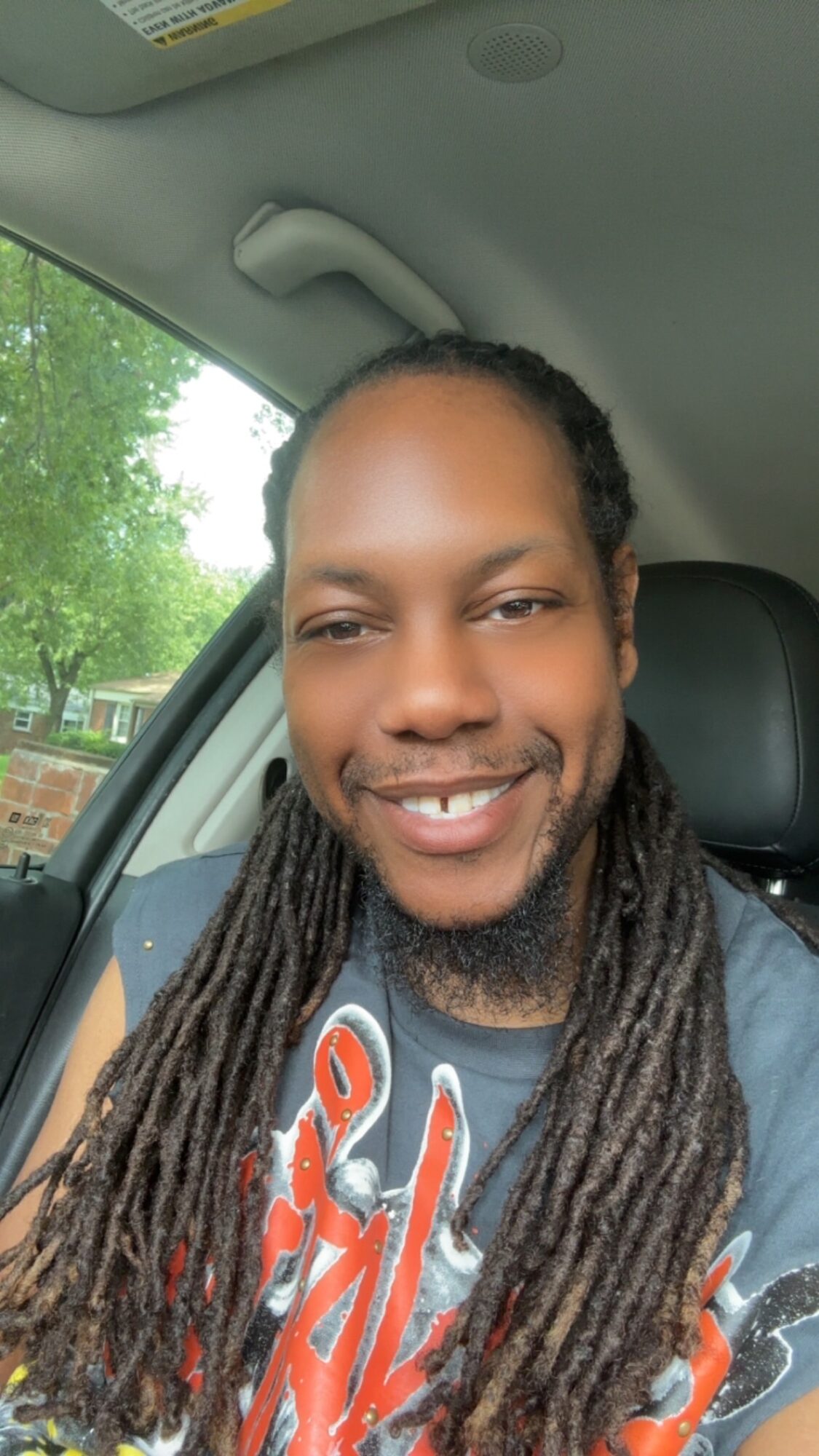 Smiling person with long dreadlocks sitting in a car, with a window showing trees outside.