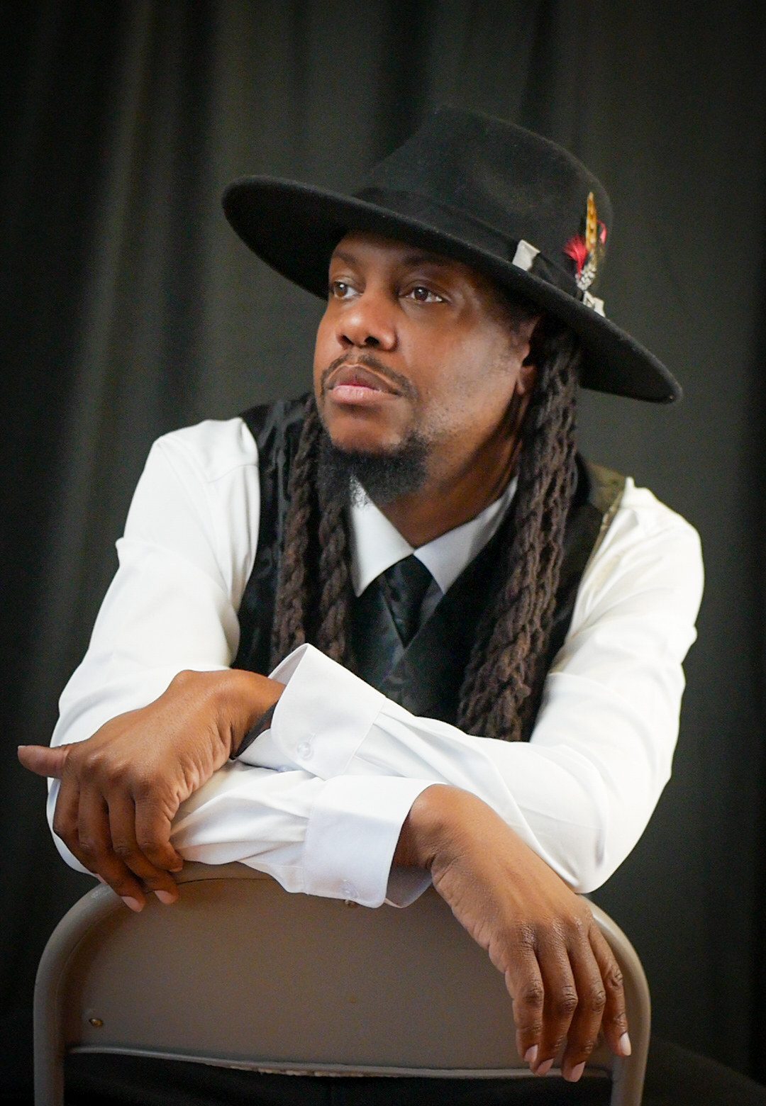 Man with long dreadlocks wearing a black hat, white shirt, and black vest, sitting with arms crossed, looking to the side.