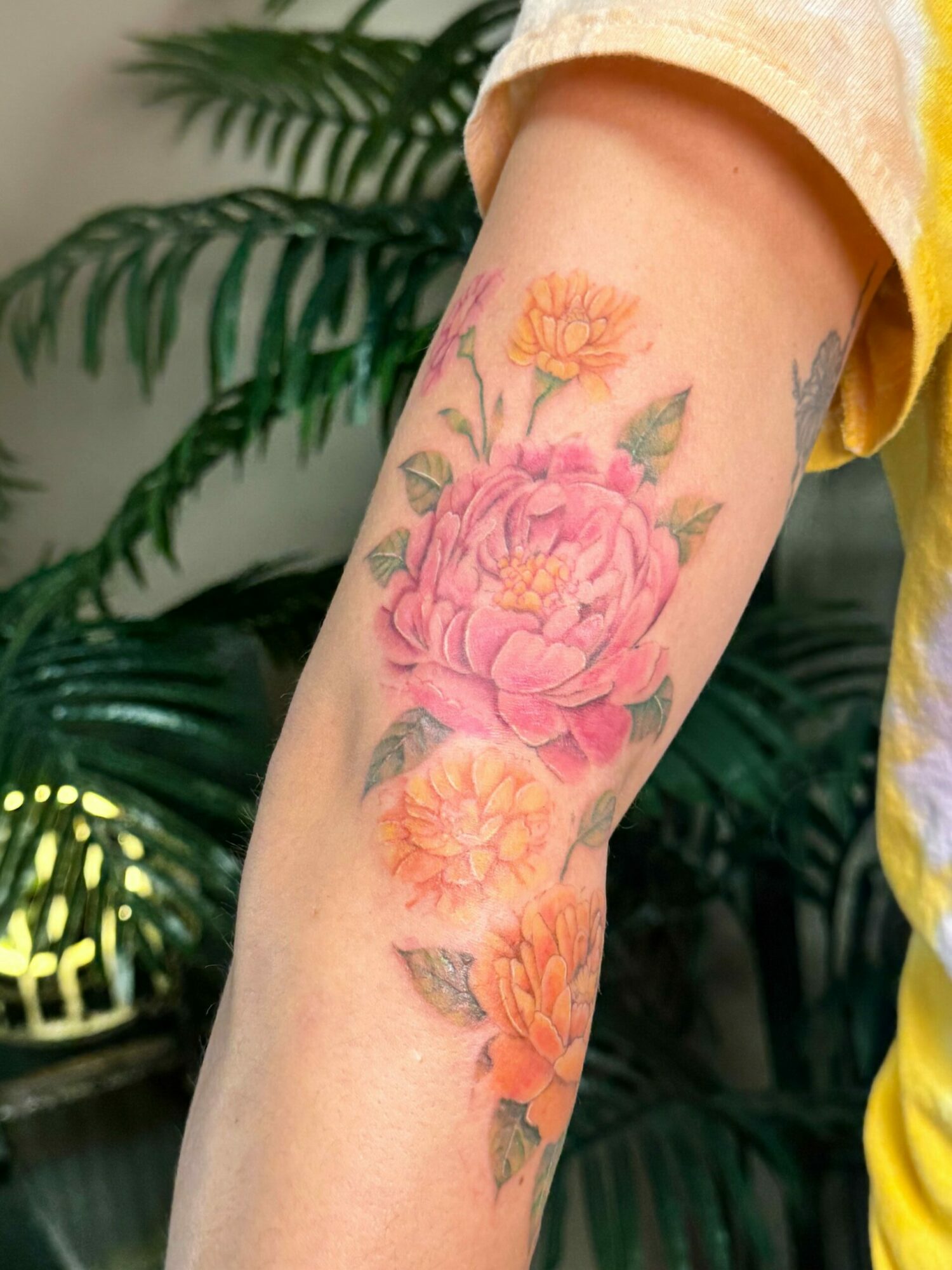 Forearm tattoo of pink and yellow flowers with green leaves, against a background of green plants.
