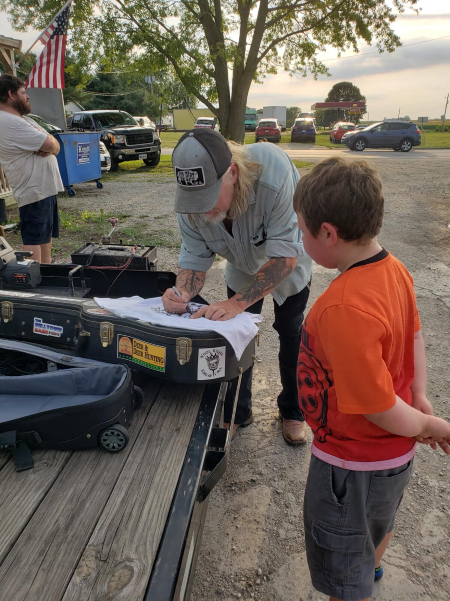 Person signing a paper on a table outdoors, with a child in an orange shirt nearby, trees and cars in background.