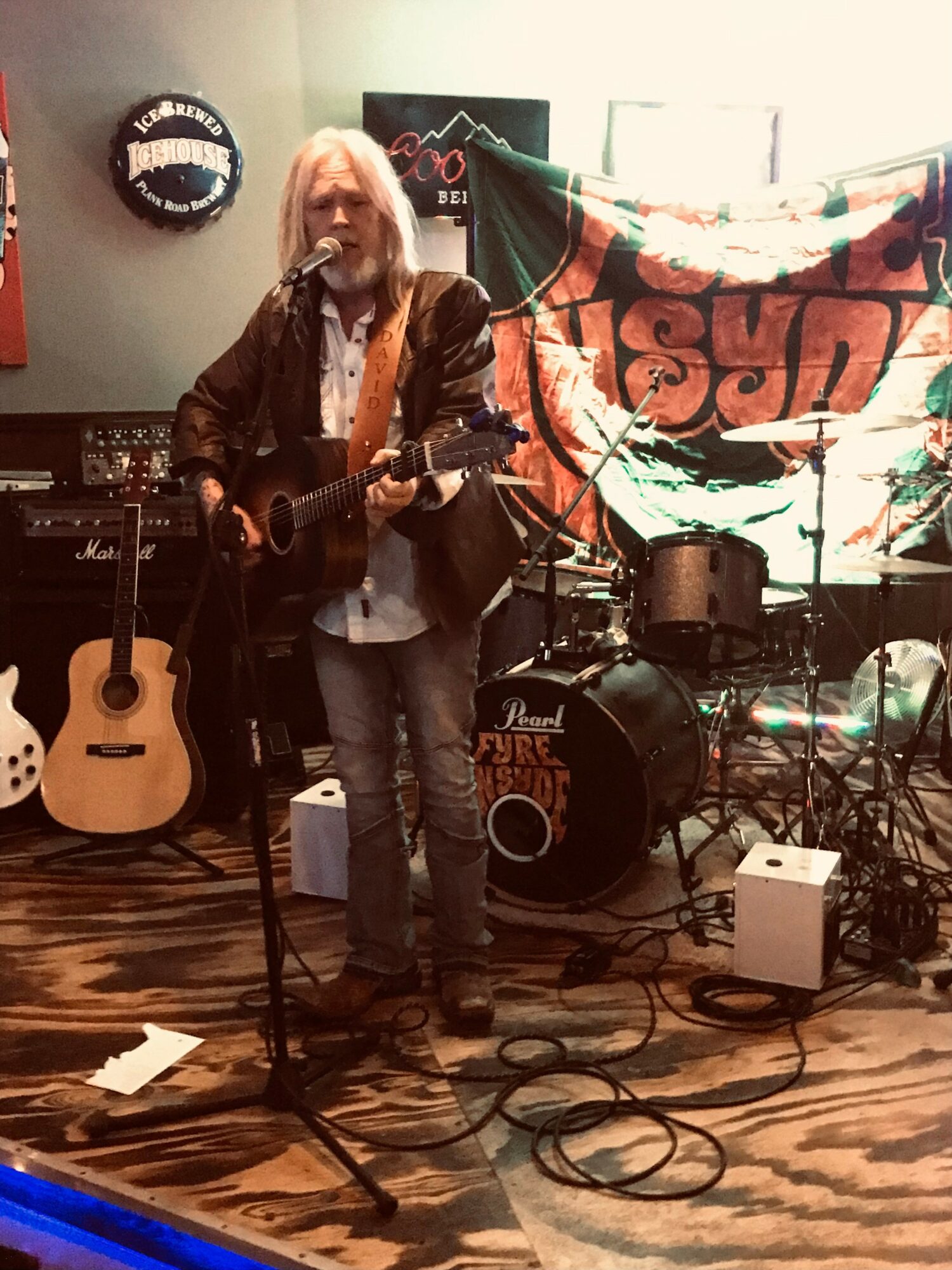 Person with long hair playing guitar on stage with drums and guitars, banner in background, indoor setting.