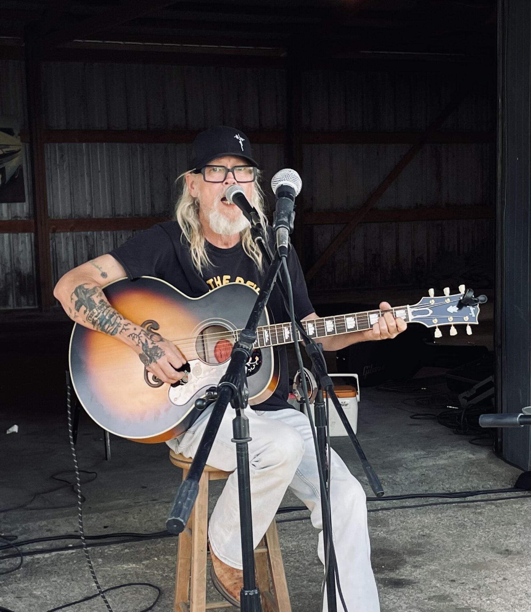 Person with long hair, glasses, and tattoos plays an acoustic guitar and sings into a microphone inside a dark, industrial space.