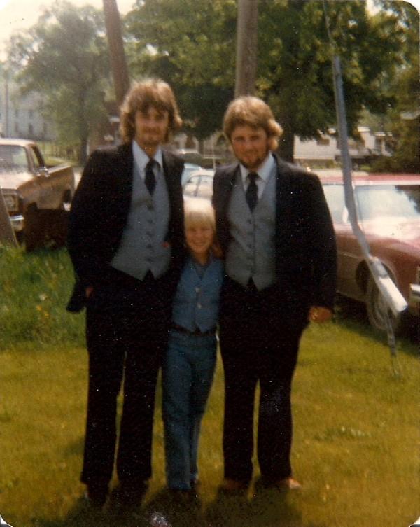 Three people standing outdoors on grass, two adults and one child, dressed in formal suits, smiling at the camera.