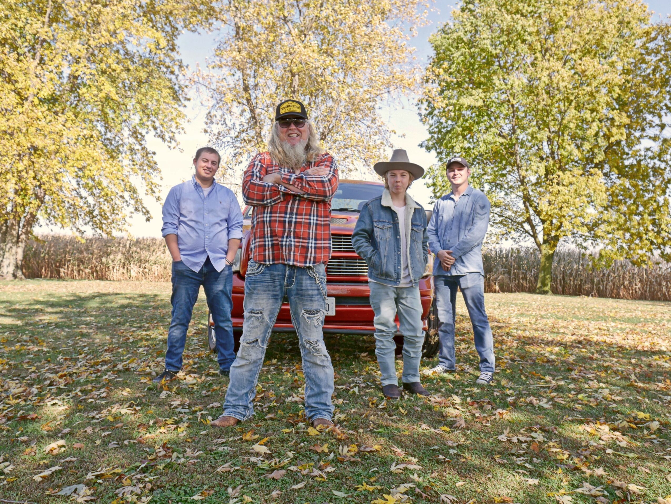 Four people standing outdoors on grass with trees and a vehicle in the background.