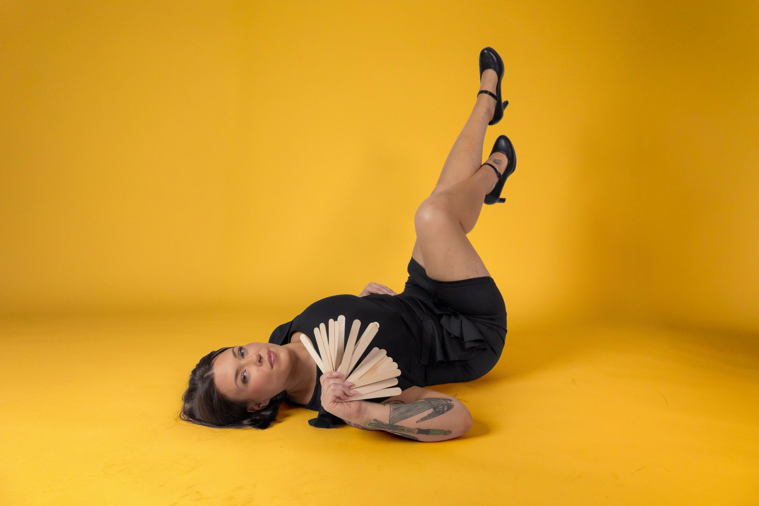 Woman lying on yellow floor holding a fan, with legs raised and crossed, against a yellow background.