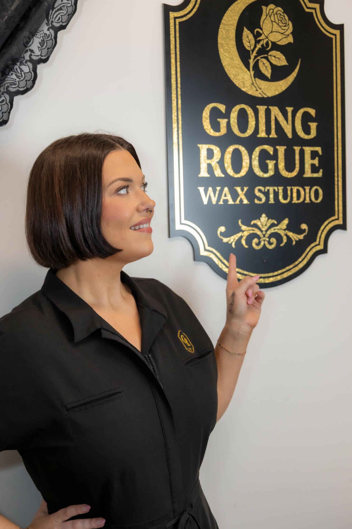 Woman with short dark hair pointing at a sign for Going Rogue Wax Studio, smiling, wearing a black uniform, indoors.