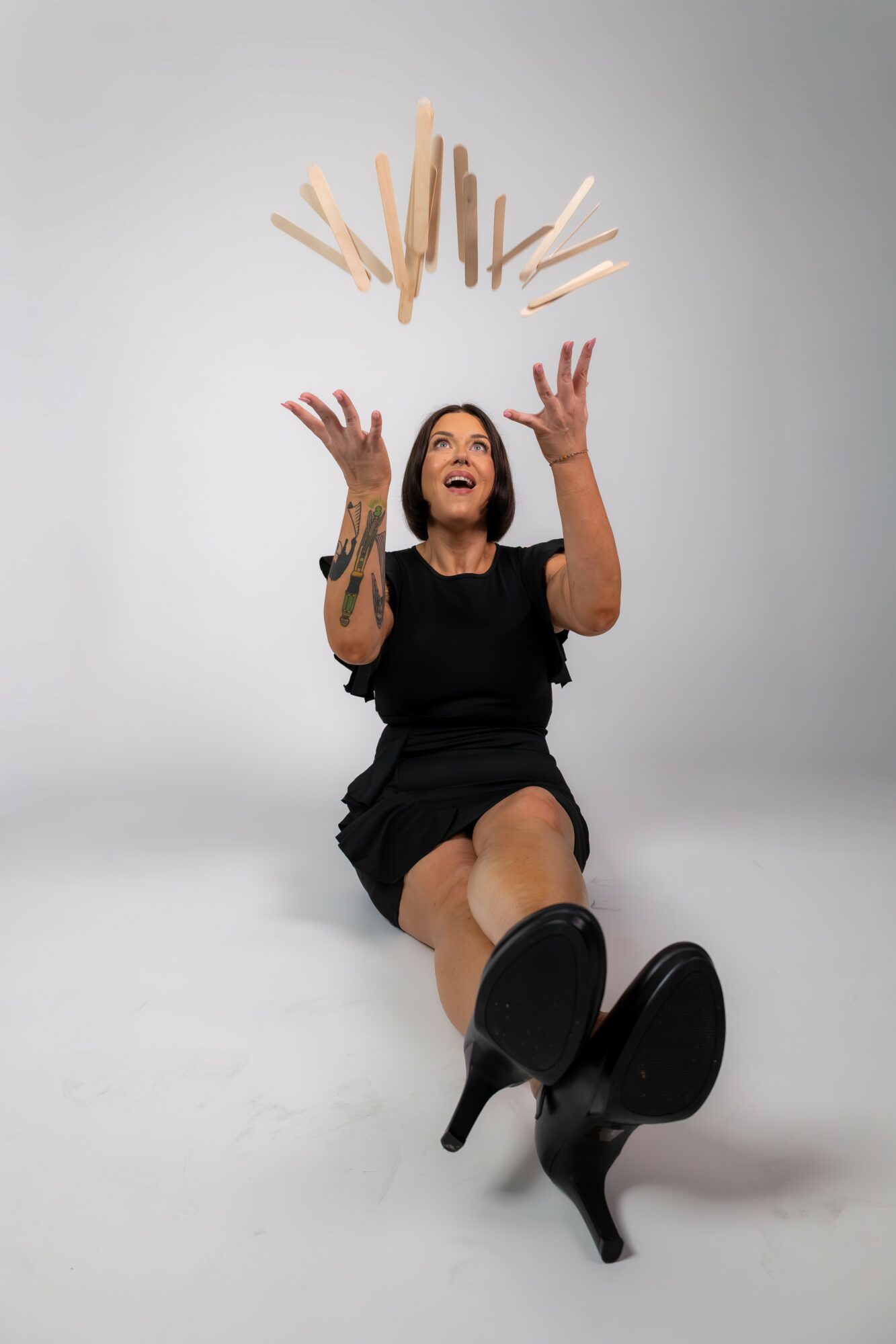 Woman sitting on the floor, smiling, with wooden sticks falling above her, wearing a black dress and high heels.