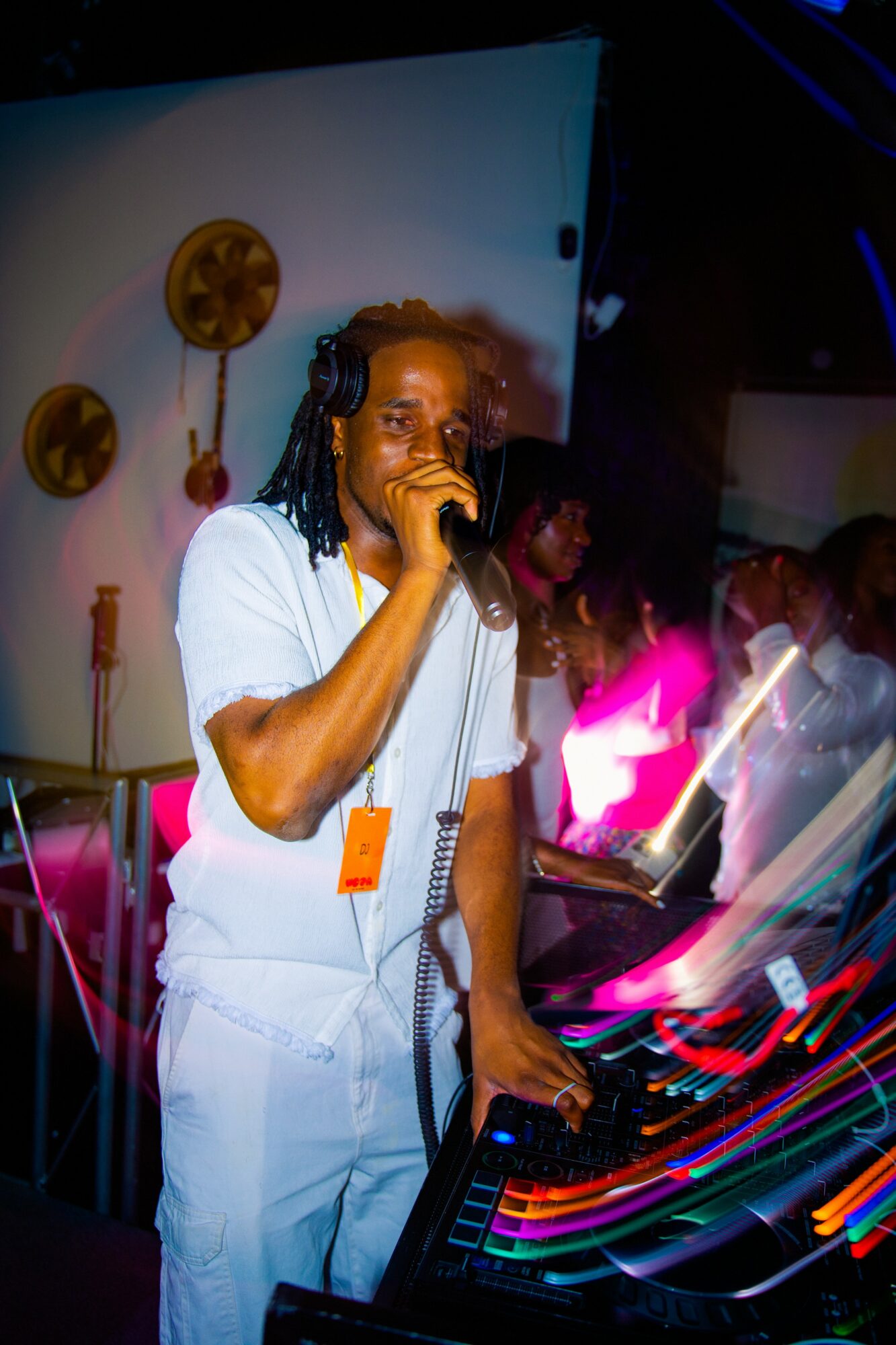 DJ with headphones and microphone performing at a party with colorful lights and people in background.