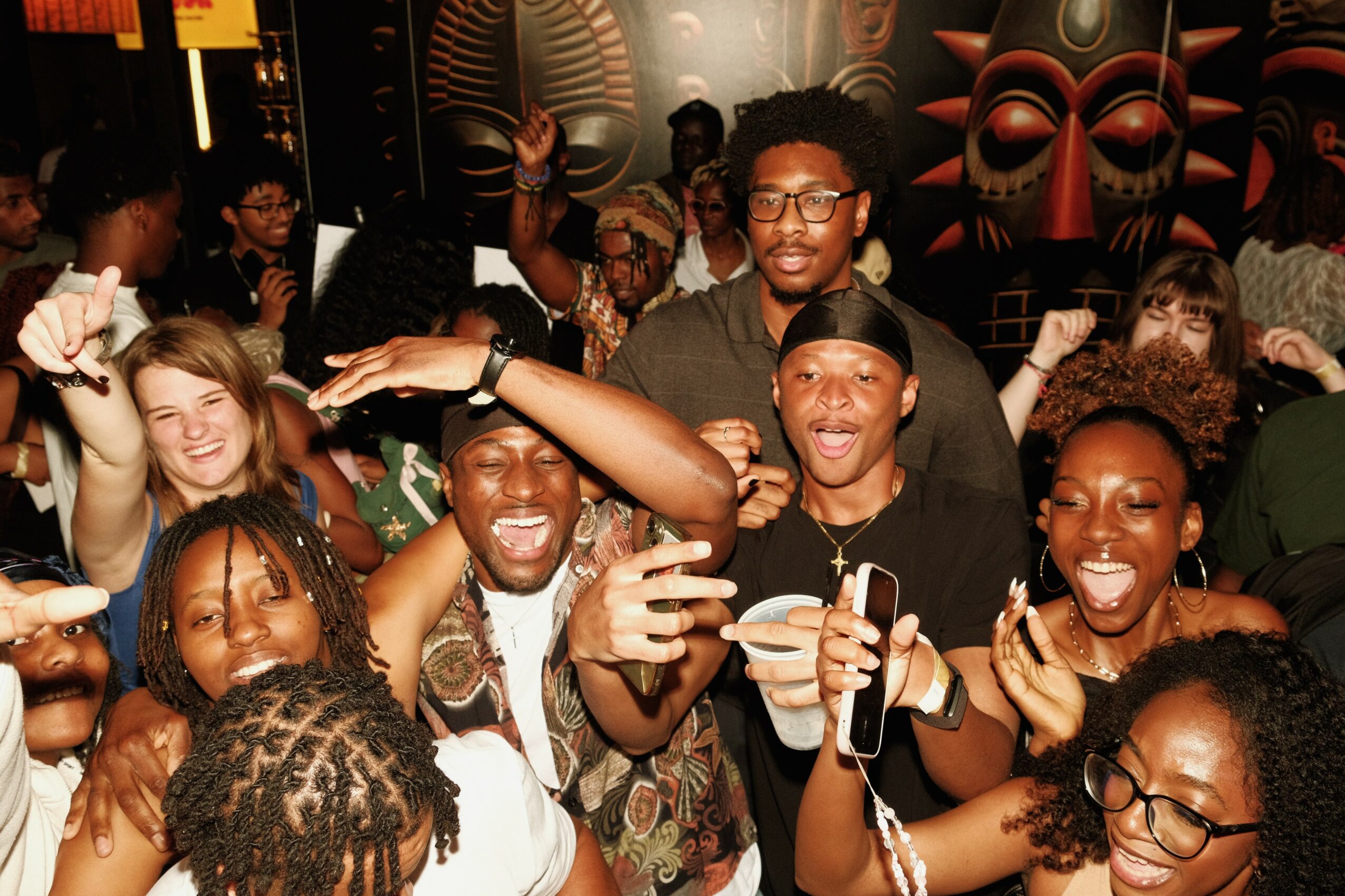 Group of people smiling and celebrating at a party, holding drinks, with colorful masks and decorations in the background.