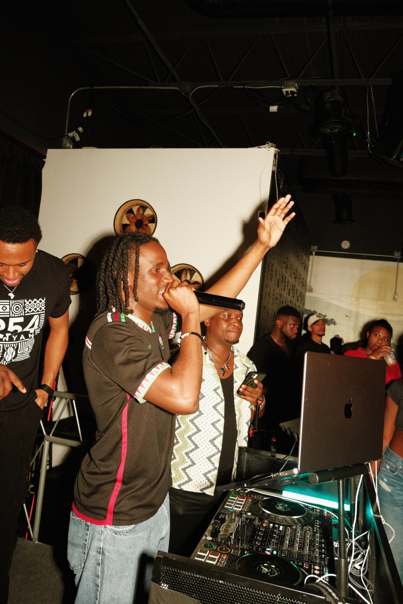 Group of people at a DJ booth with a person raising hand, in a dark room with a white backdrop.