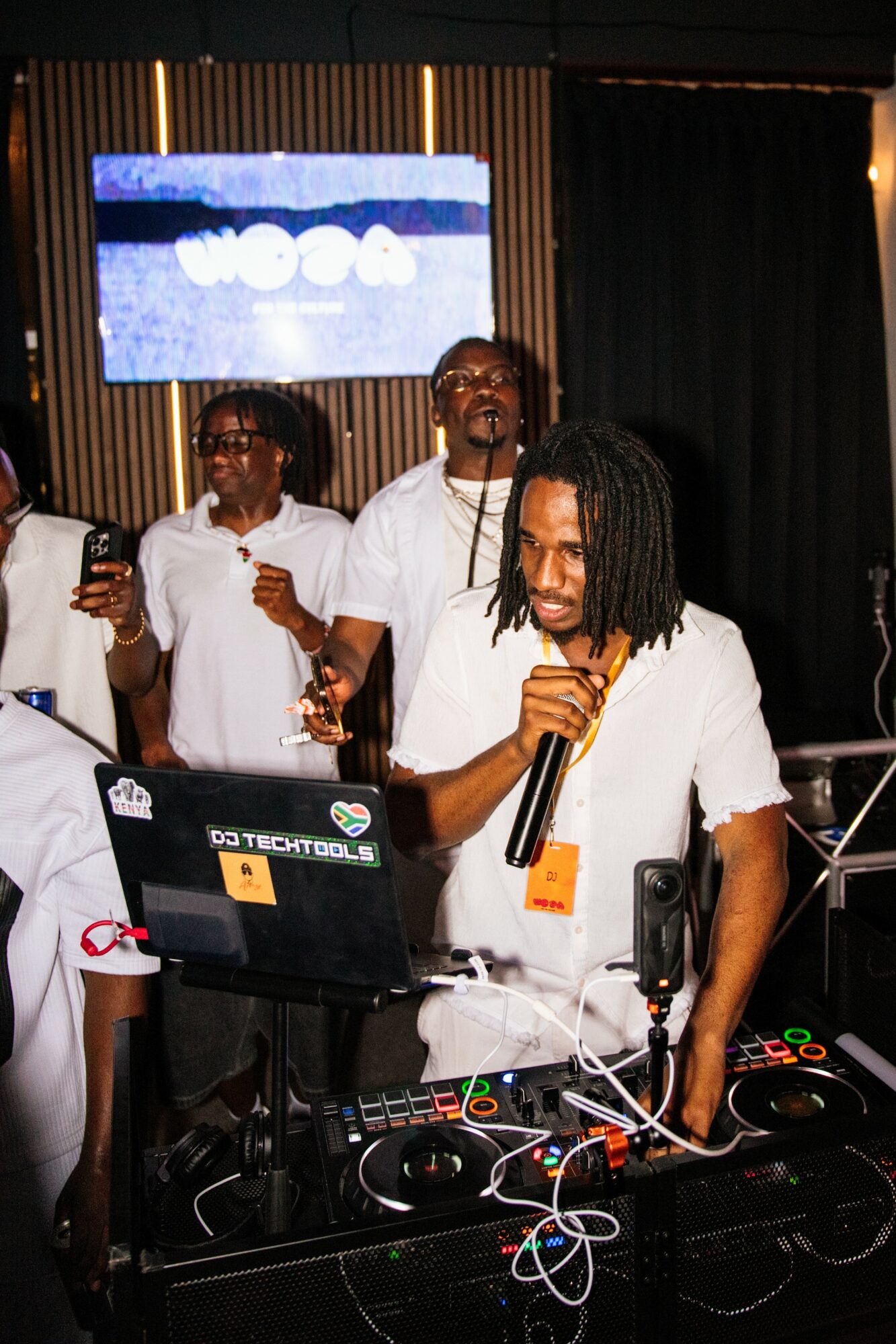 Group of people at a DJ booth, one person with dreadlocks holding a microphone, others smiling and dancing, screen in background