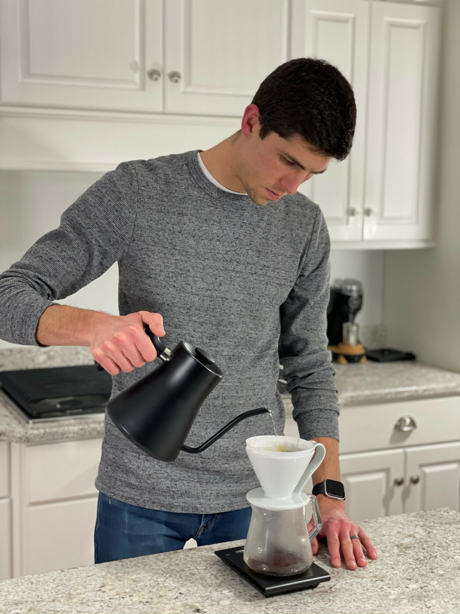 Young man pouring hot water into a coffee dripper on a kitchen counter, with white cabinets in the background.