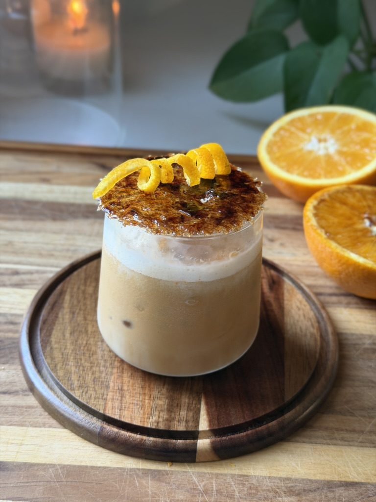 A creamy beverage topped with caramelized sugar and lemon peel, served in a glass on a wooden tray with oranges and green leaves in the background.