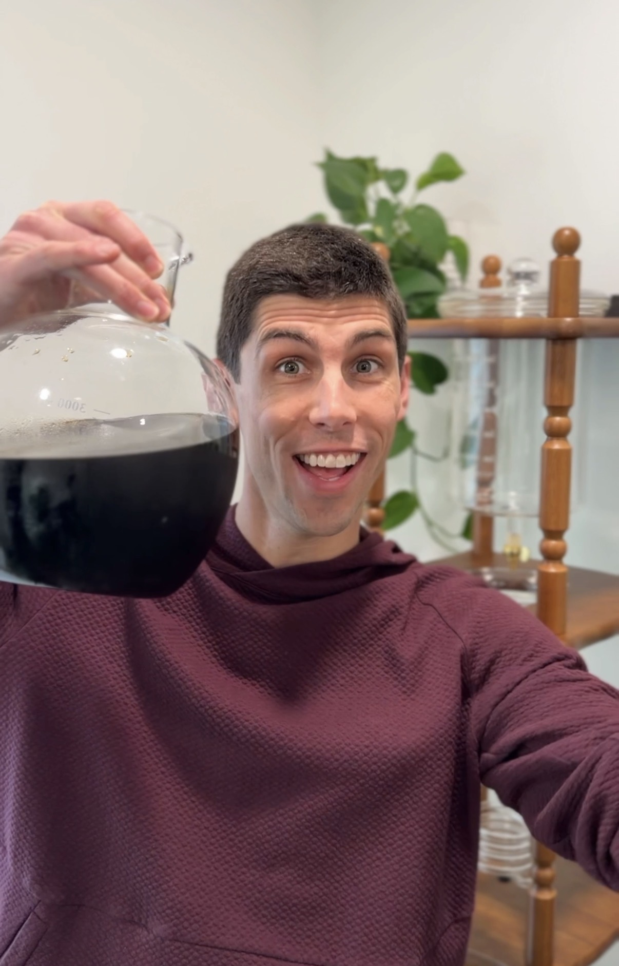 Person taking a selfie, holding a glass container with dark liquid, smiling, with a plant and shelves in background.