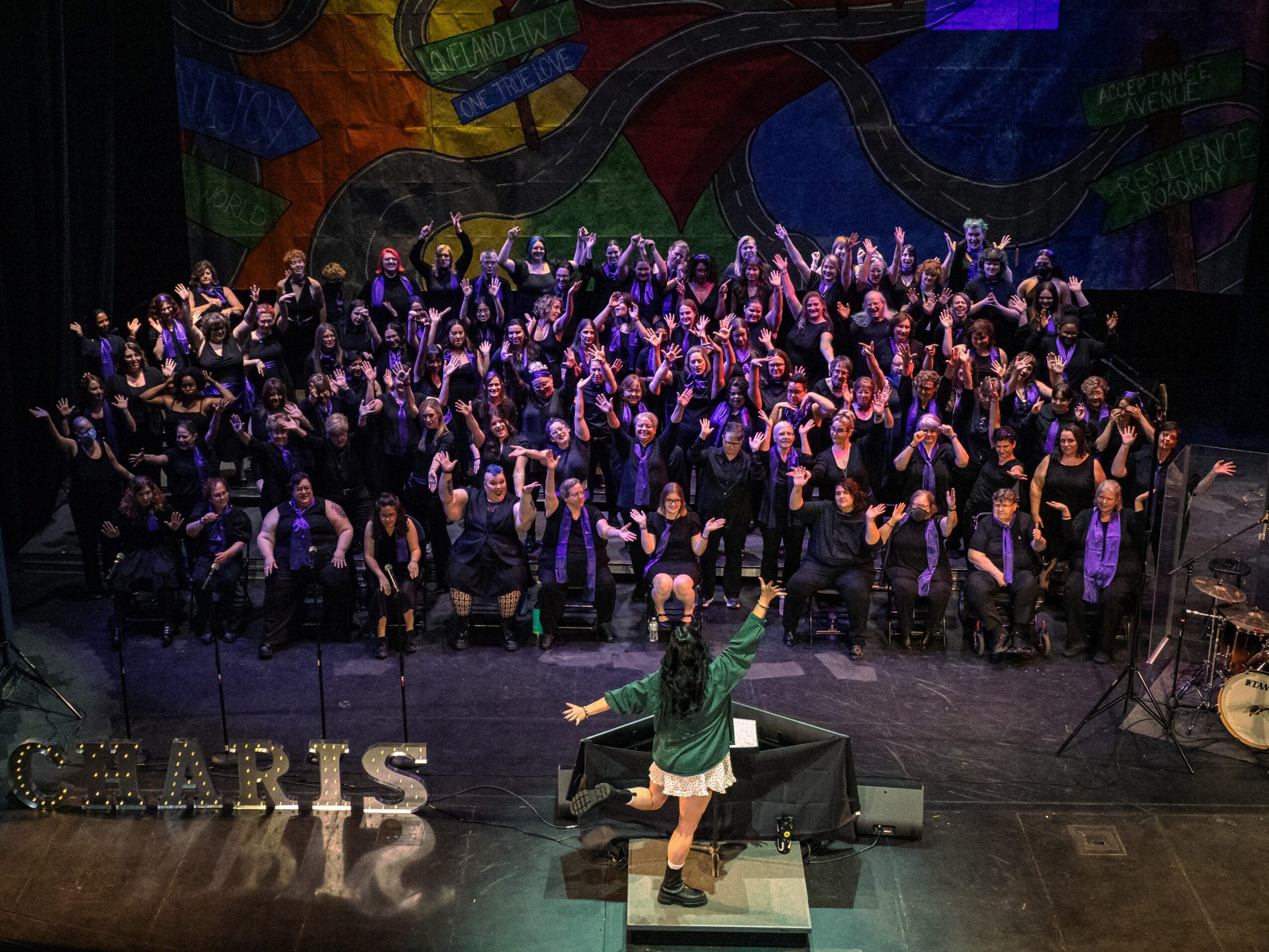 Large group of people on stage with a colorful backdrop, some raising hands, a person in front conducting, and musical instruments nearby.