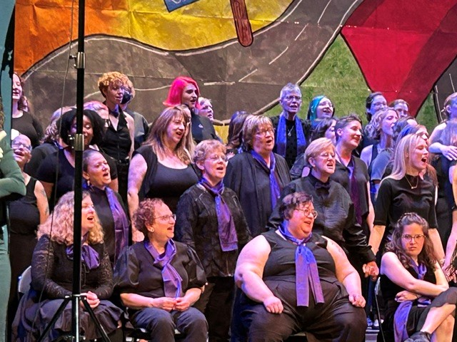 Group of children and adults singing on stage with colorful background, some wearing black shirts, smiling and looking forward.