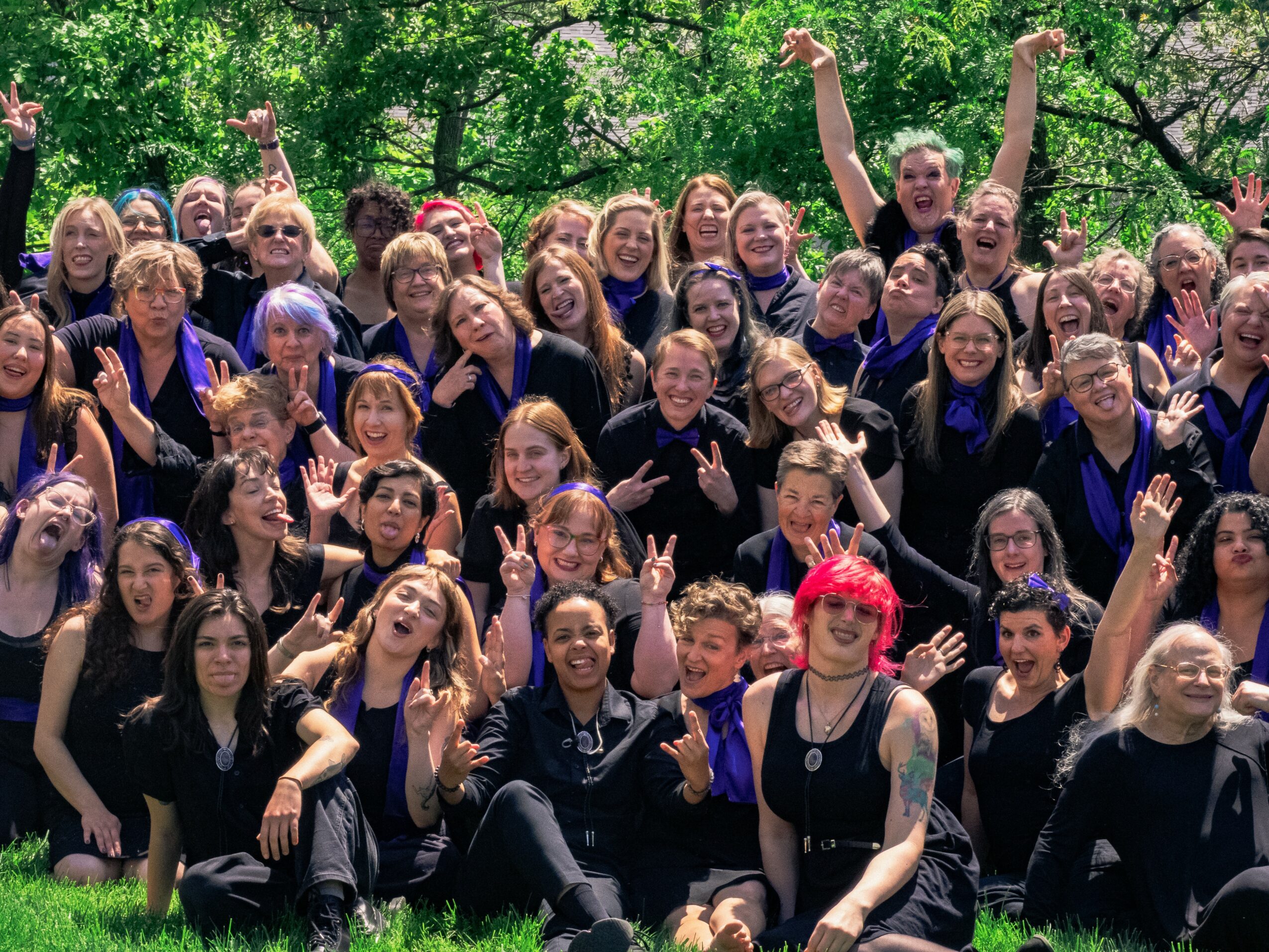 Group of smiling women and girls outdoors, some making peace signs, in a green park with trees.