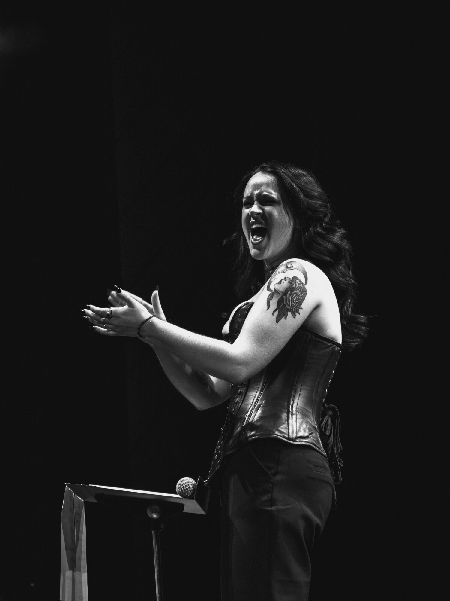 A woman with long dark hair, wearing a corset, is laughing and clapping her hands on stage.
