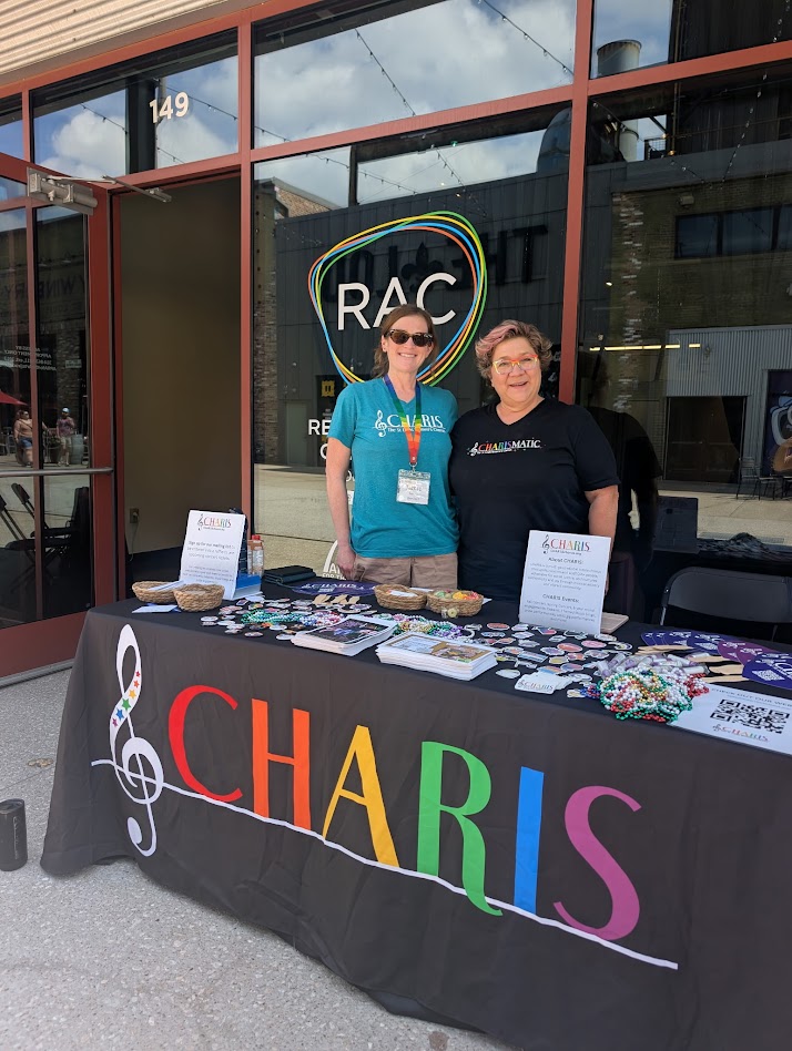 Two women stand behind a table with colorful CHARIS letters, brochures, and items outside a building with RAC logo.