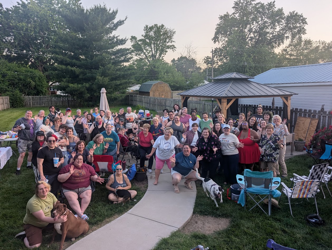 Group of people gathered outdoors in a backyard, some sitting and some standing, with trees and structures in the background.