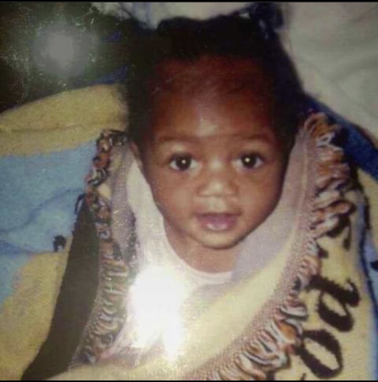 Young child with dark hair and big eyes, lying on a blanket with colorful patterns, looking at the camera.