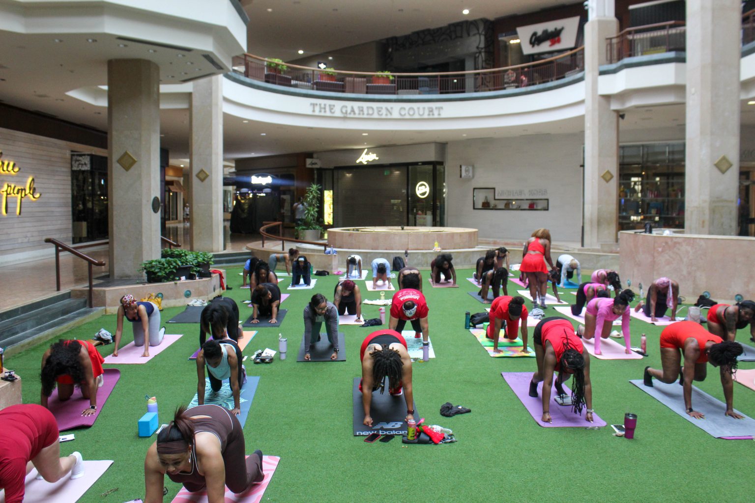 Group of people practicing yoga on green mats in a shopping mall's open area, with stores and balconies visible above.