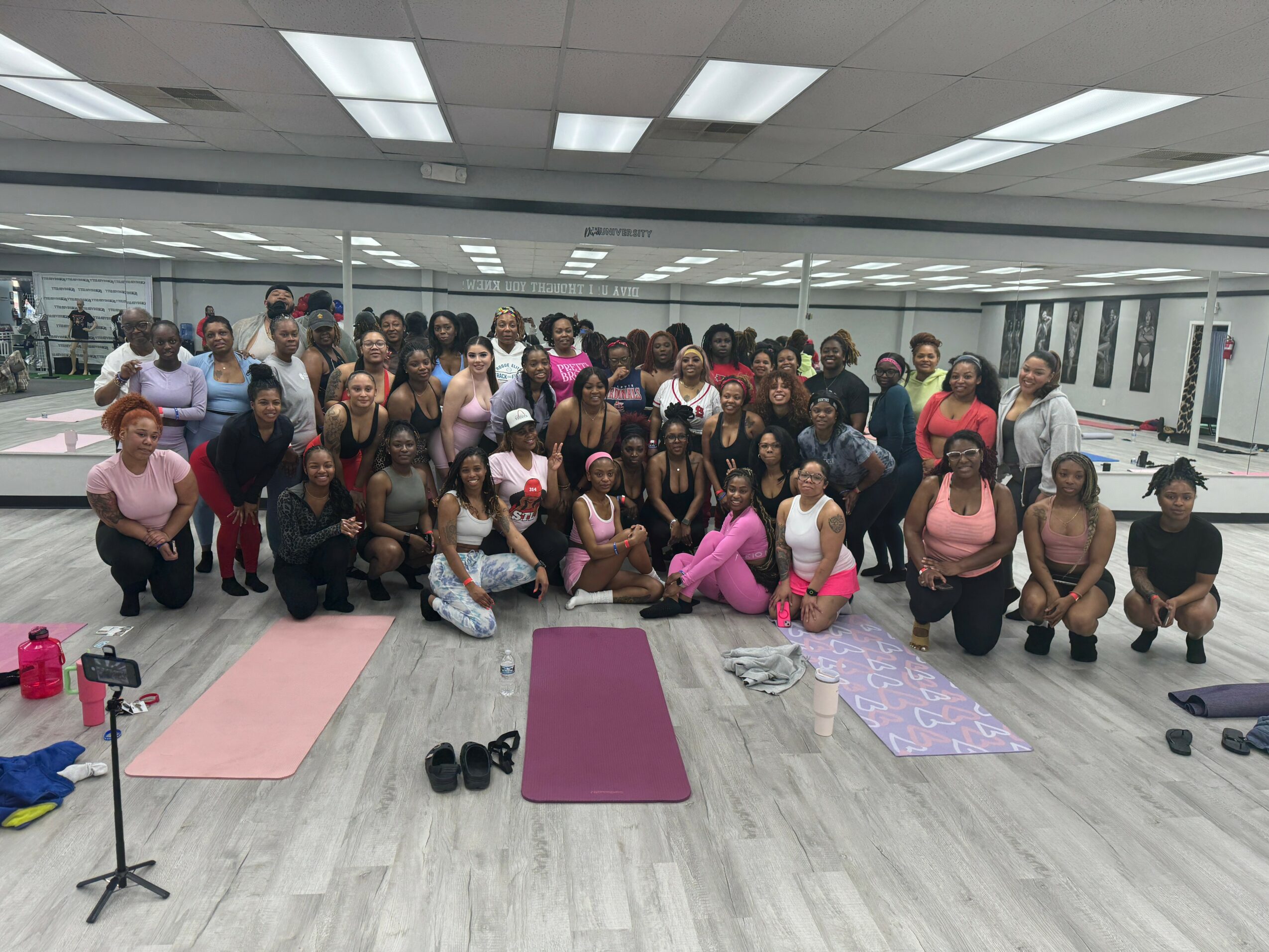 Group of people in a fitness studio with yoga mats, some kneeling and some standing, smiling at the camera.