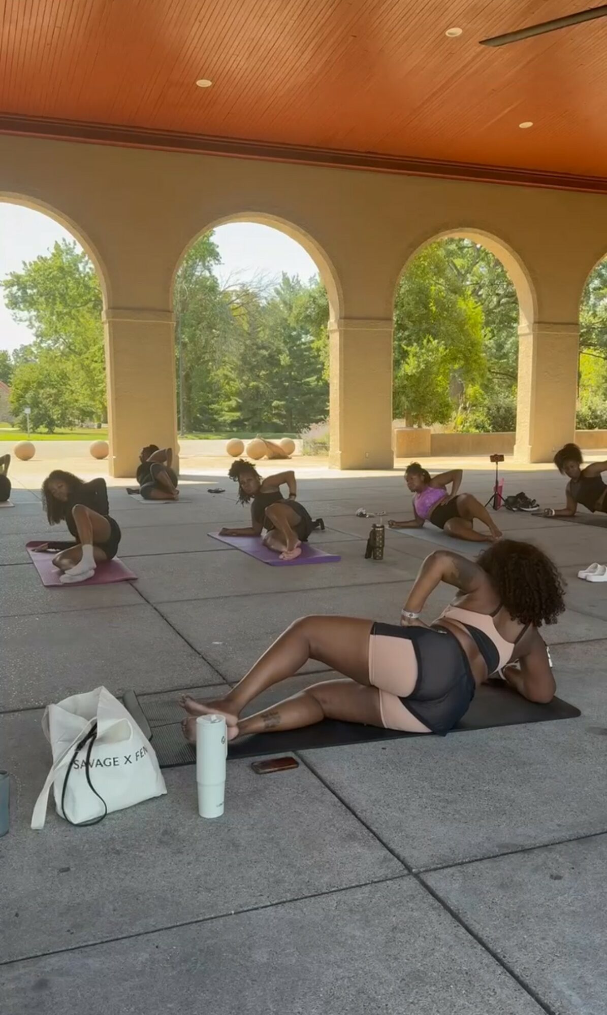 Group of people practicing yoga outdoors on mats under arches, with trees and sky visible in the background.