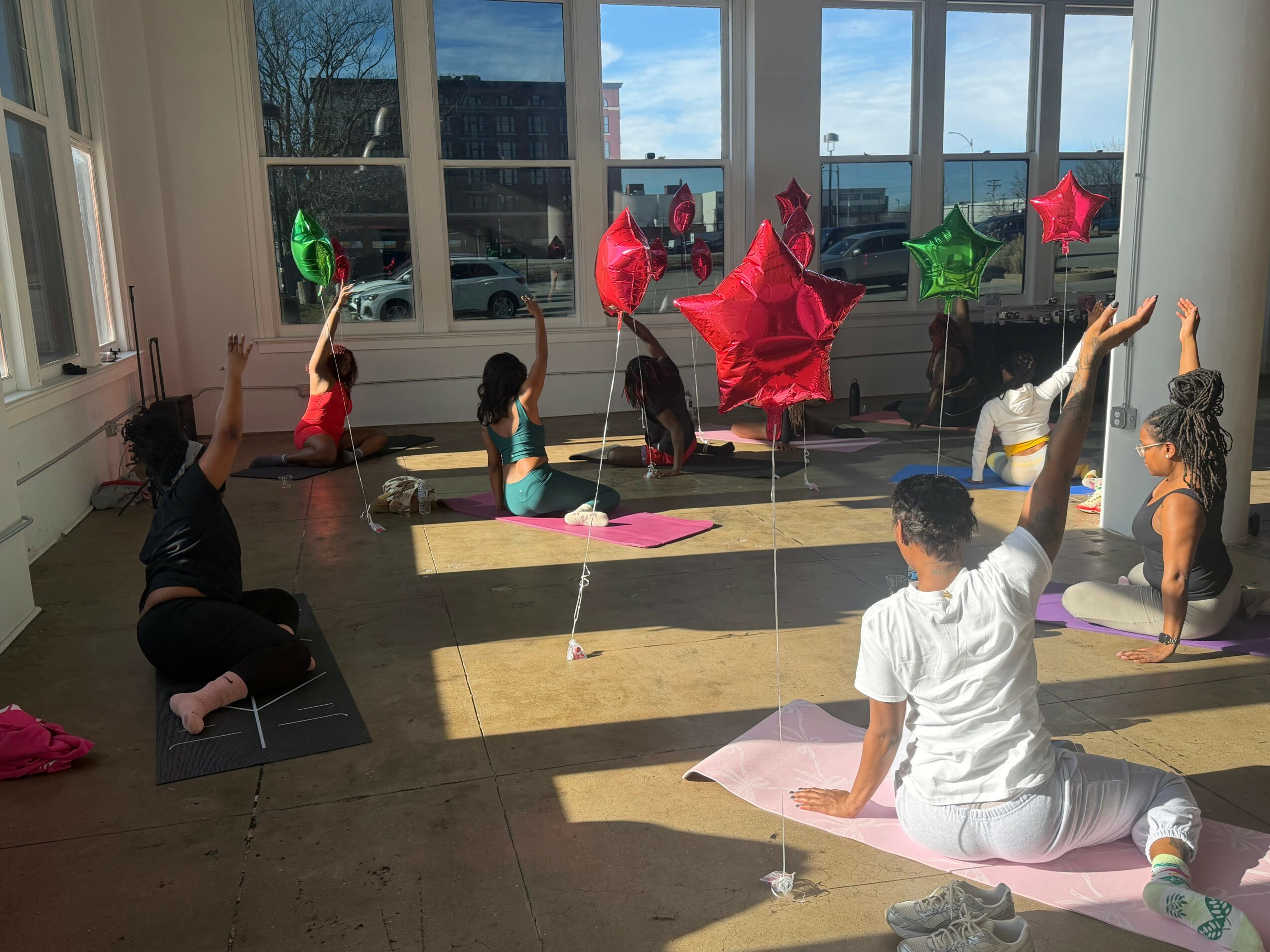 People practicing yoga in a bright room with large windows, colorful balloons, and sunlight streaming in.