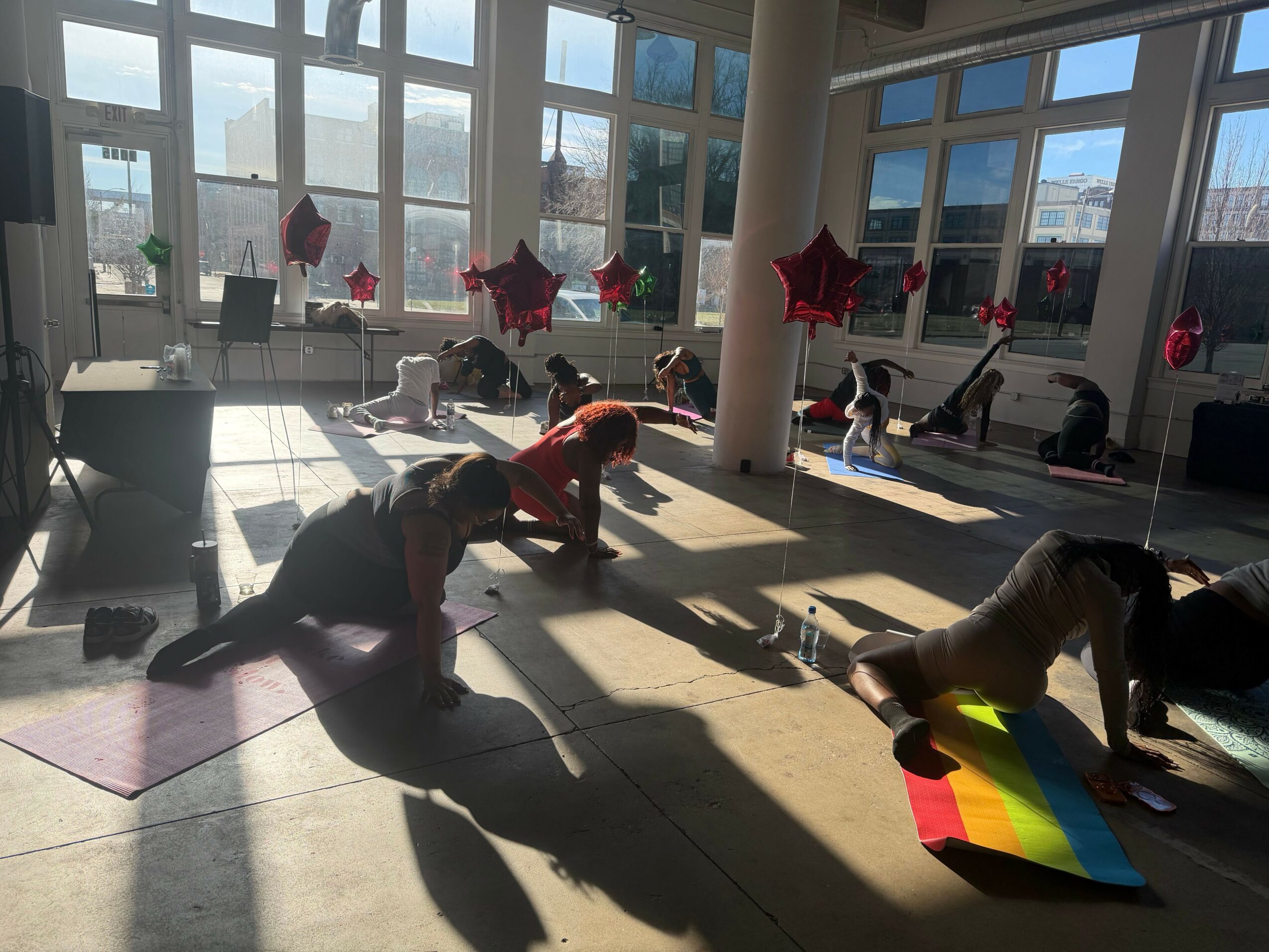 People are dancing and playing with a rainbow-colored cube in a room with large windows and red star-shaped balloons.