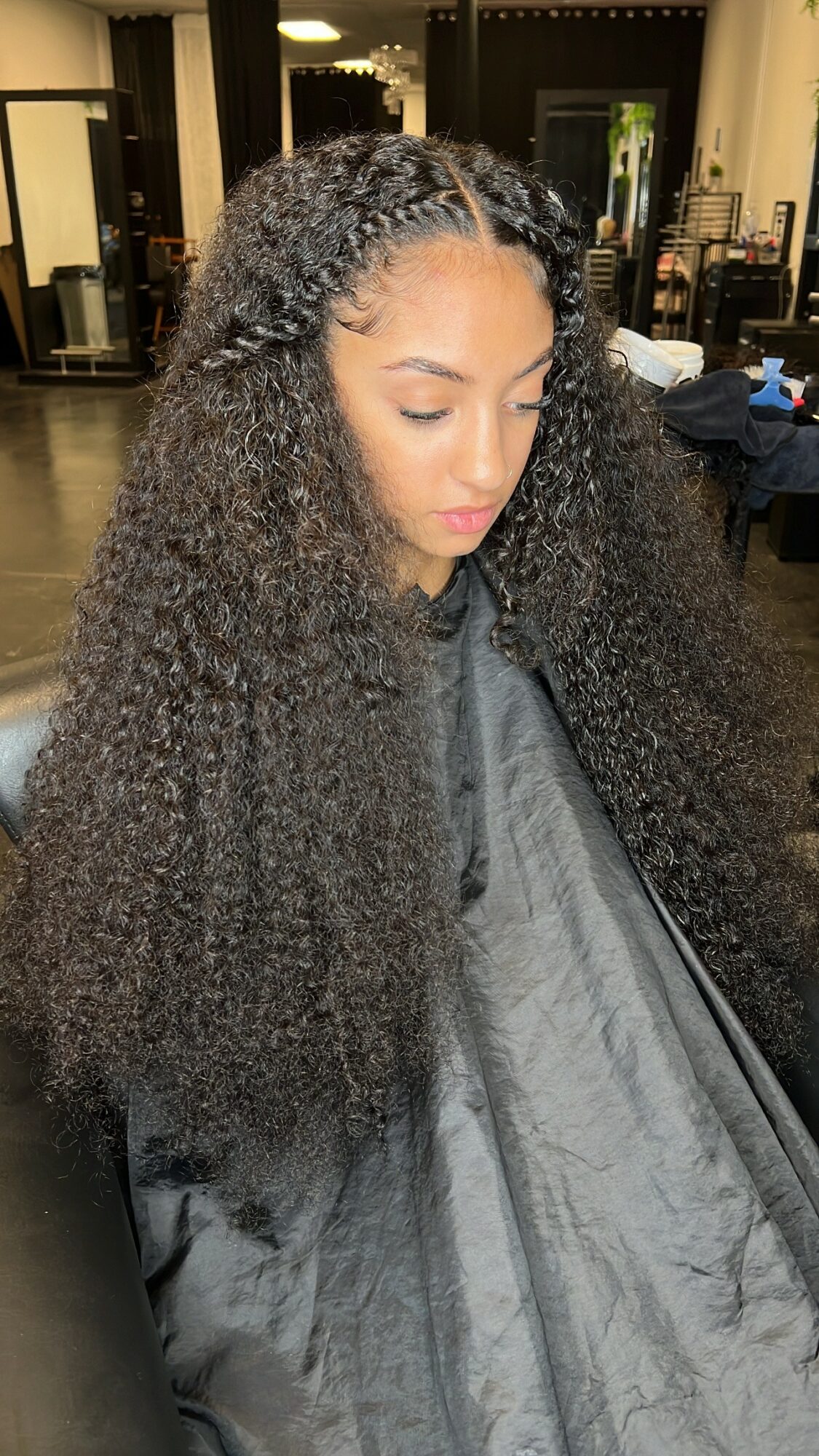 Young woman with long, curly hair looking down in a salon, wearing a cape, with salon equipment in background.