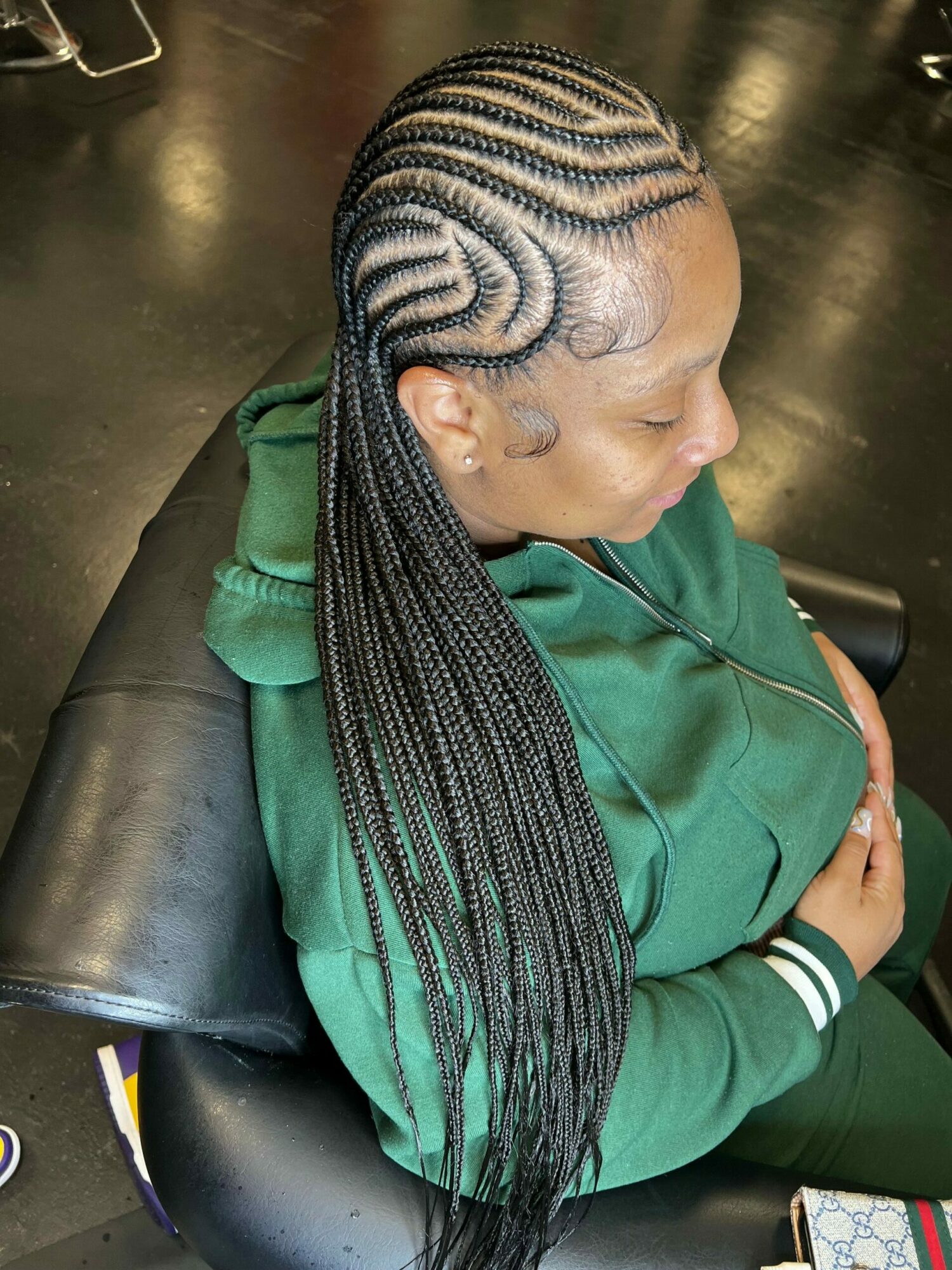 Person with long braided hair and patterned scalp, wearing a green jacket, sitting on a black chair with hands folded.