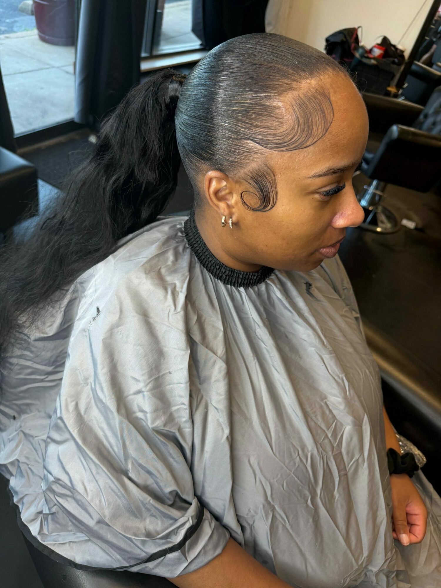 Side view of a woman with long black hair, wearing a gray cape, sitting in a salon chair.