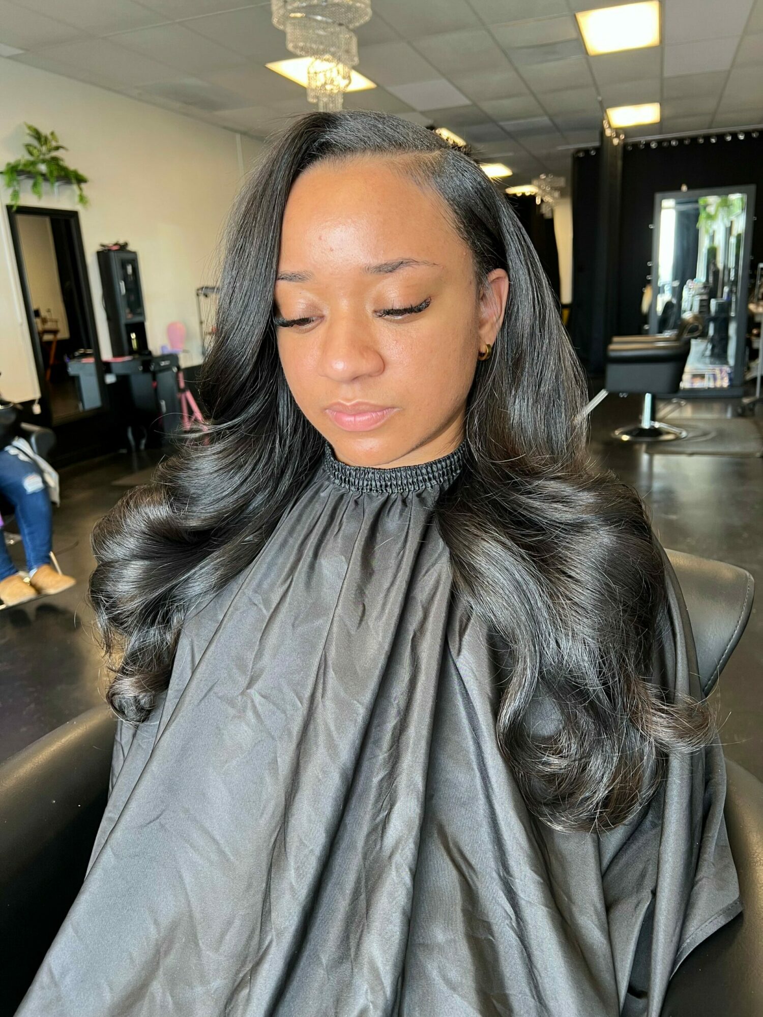 Woman with long black hair and closed eyes, sitting in a salon chair with a cape, in a modern salon interior.