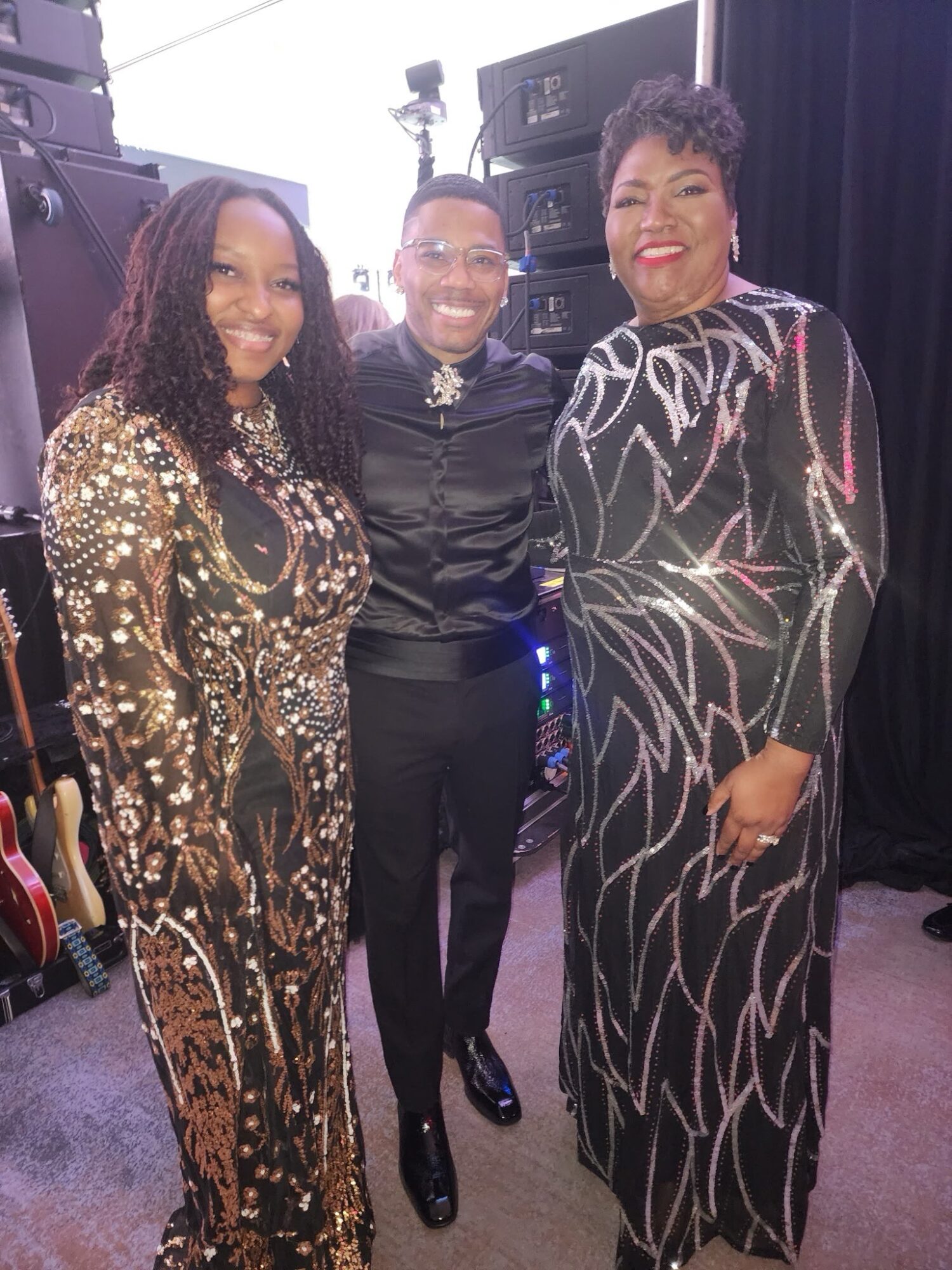 Three women standing together, smiling, in a studio or stage setting with equipment in the background.