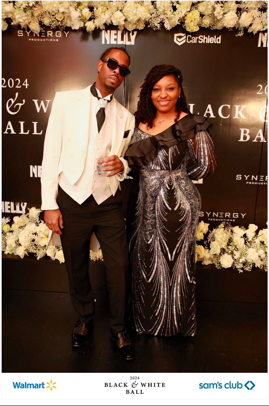 A man and woman in formal attire pose together on a red carpet with a black backdrop and floral decorations.
