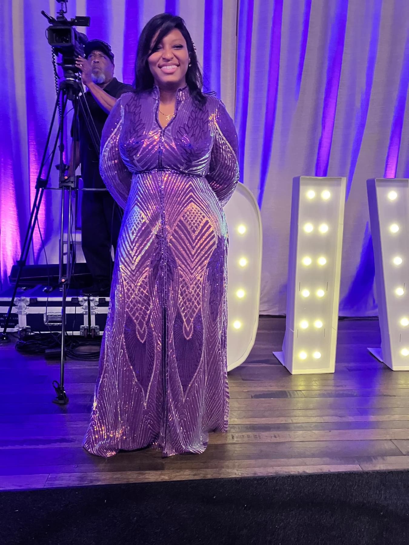 Woman in a purple, sequined gown standing on stage with large illuminated letters behind her.