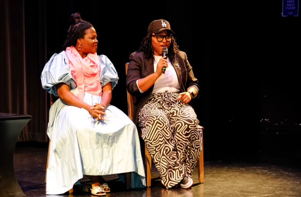Two women sit on stage, one speaking into a microphone, the other listening, with a dark background.